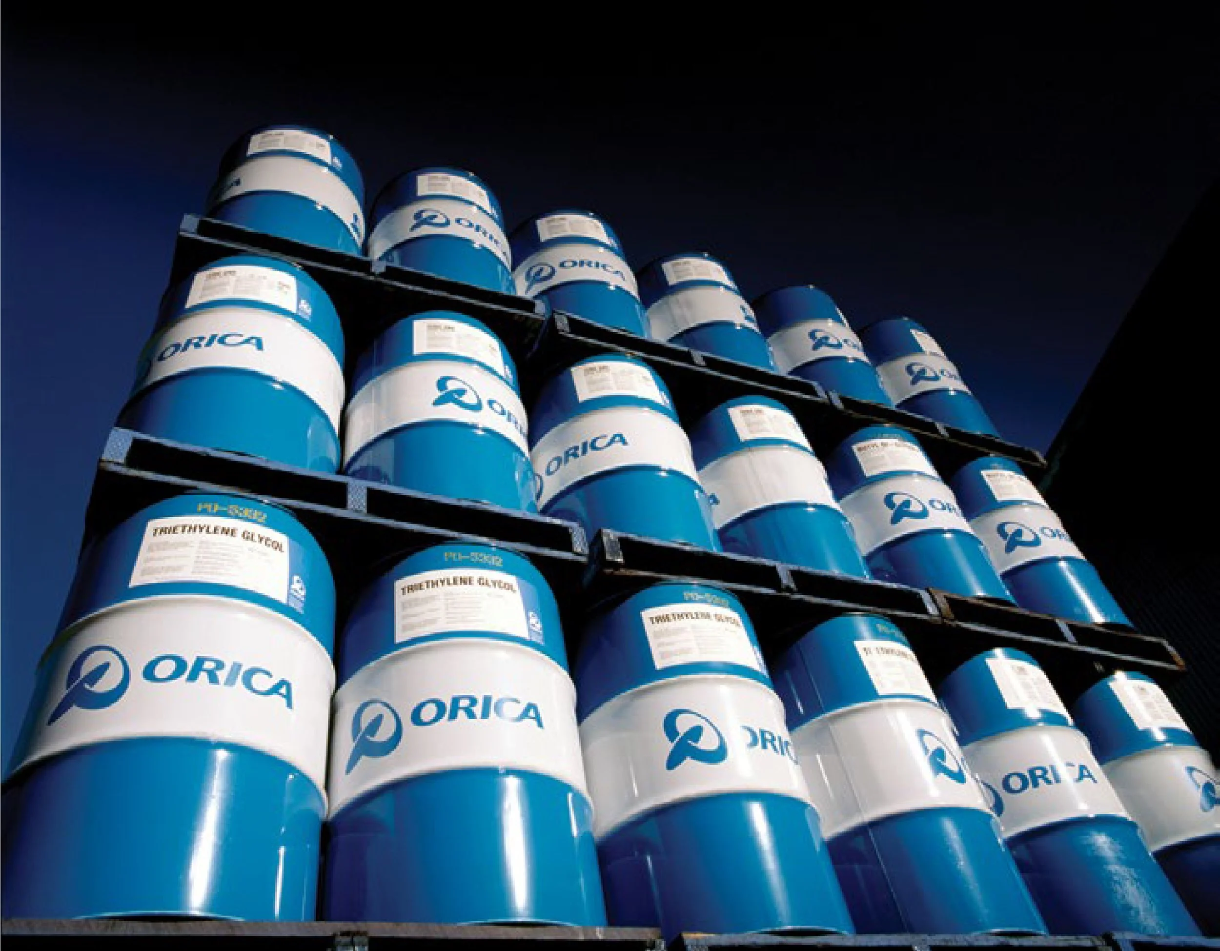 Multiple blue and white barrels stacked on shelves with the logo of Orica, a chemical company, visible on them.