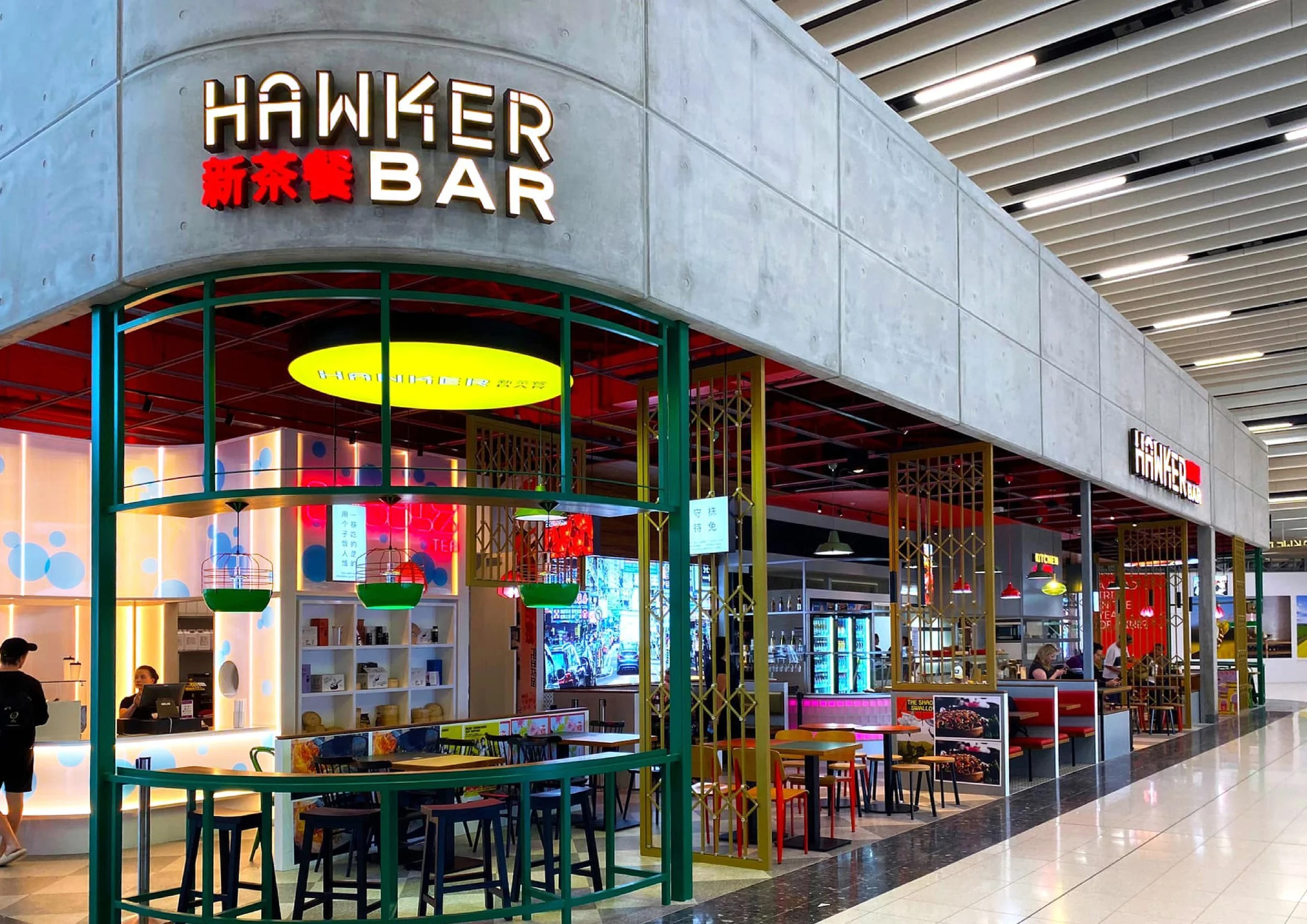 A food court area inside a shopping mall with a restaurant called 'Hawker Bar' in the background. The restaurant has a modern design with colorful decor, including hanging green and red lamps, and a curved seating area with blue chairs.