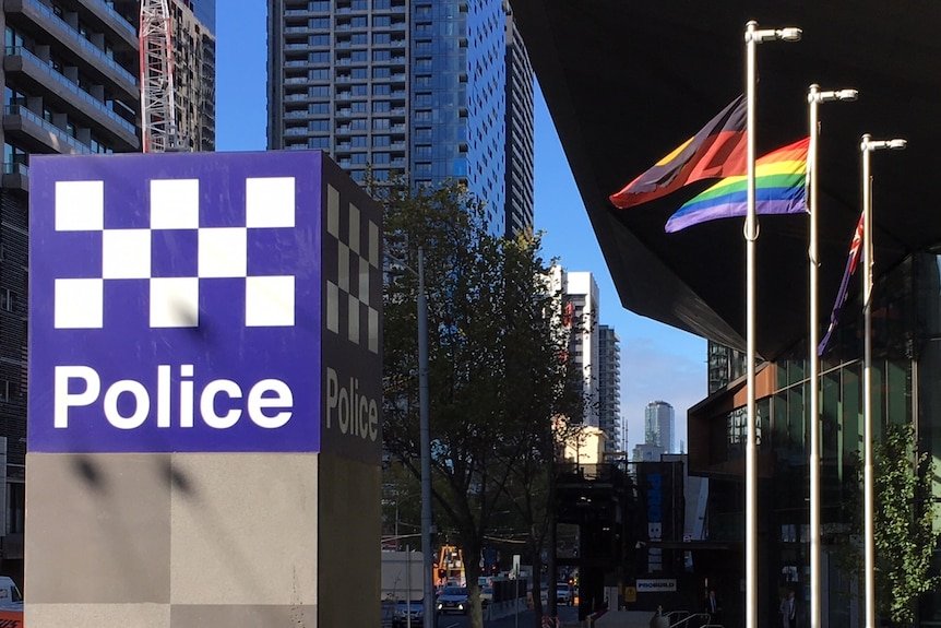 A city street scene featuring a police station sign with a checkered pattern and the word 'Police,' alongside a building with rainbow pride flags waving in the wind.