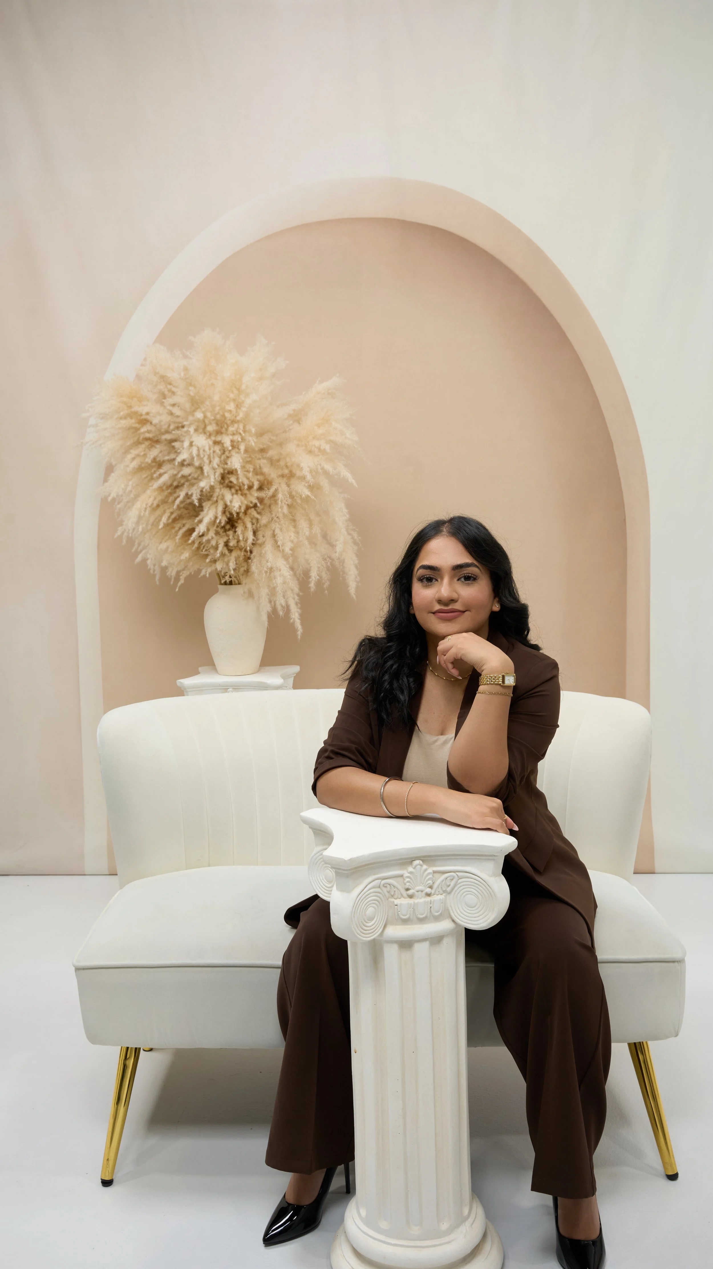 A woman with dark, wavy hair sitting on a white couch, wearing a brown blazer and matching brown pants, resting her chin on her hand, with a decorative white table and Greek column in front of her, and a large beige floral arrangement in the background.