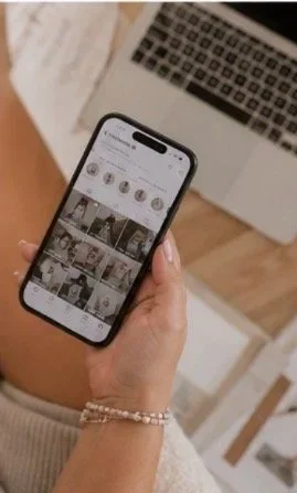 Person holding a smartphone showing a social media photo grid, with a laptop in the background on a wooden table.