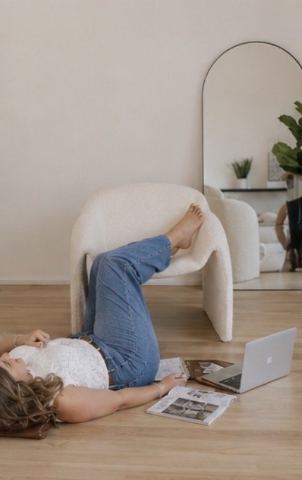 Person lying on the floor with their legs up on a white armchair, using a laptop, with a magazine on the floor nearby in a modern room with a large mirror and green plants.