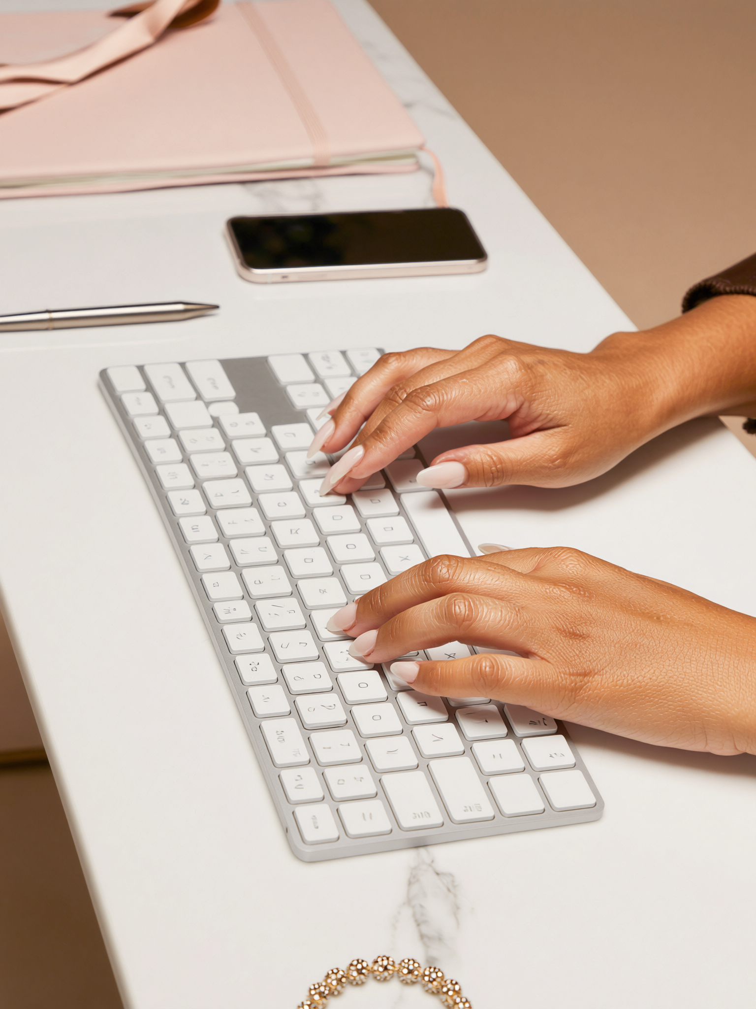 Hands typing on a white keyboard on a white table. Nearby are a pen, a smartphone, and a pink notebook with a ribbon. There is also a gold and white bracelet at the bottom of the image.