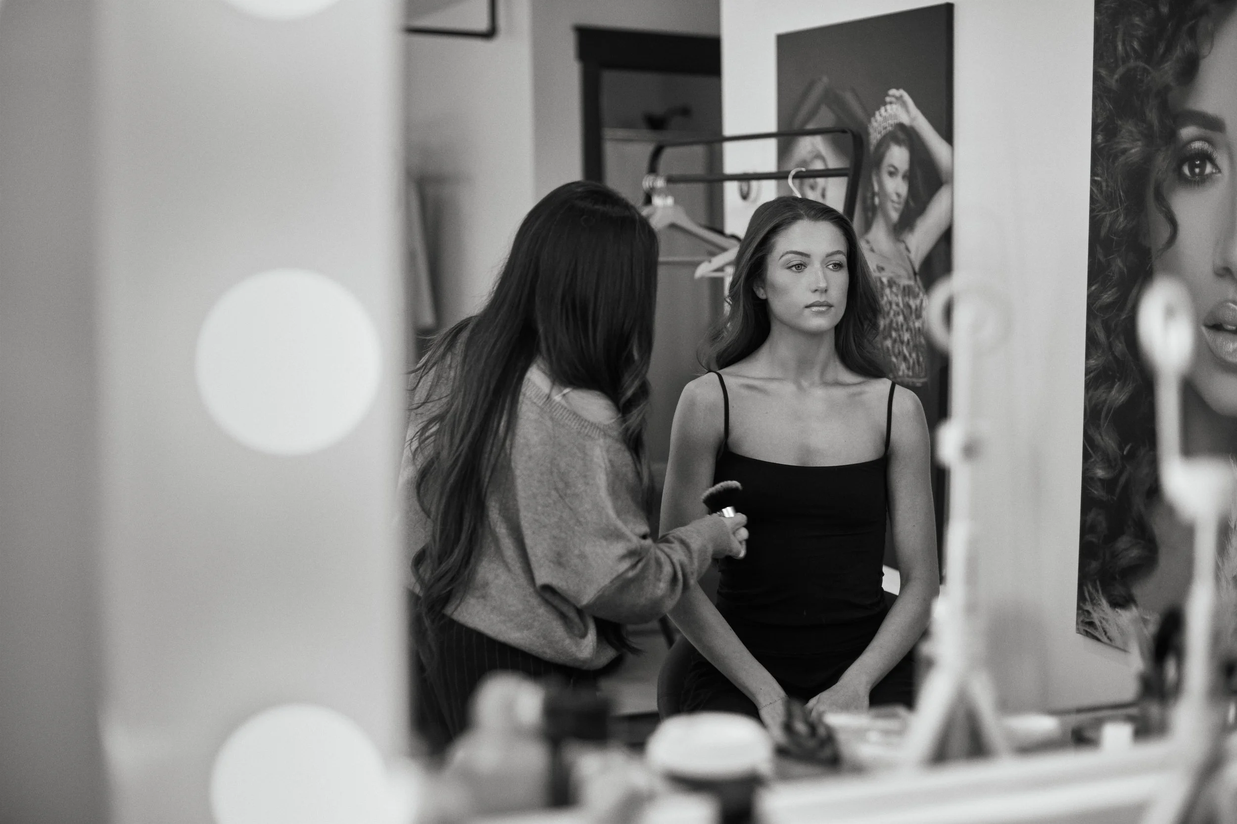 A woman with dark wavy hair getting her makeup done by a makeup artist in front of a mirror, with posters of models on the wall behind.