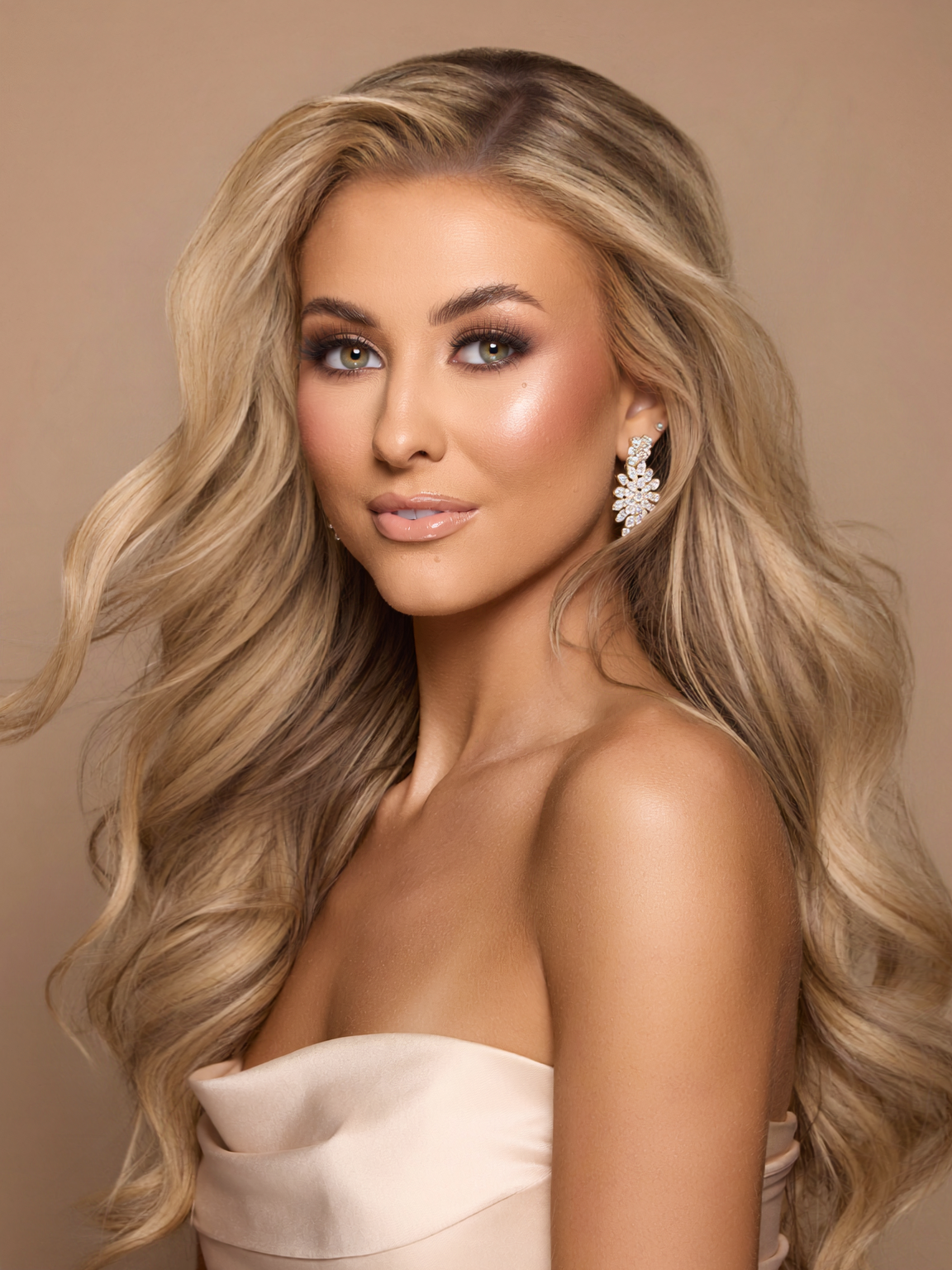 A woman with long, wavy blonde hair, wearing large, diamond-studded earrings, makeup, and an off-shoulder cream-colored dress, looking directly at the camera.