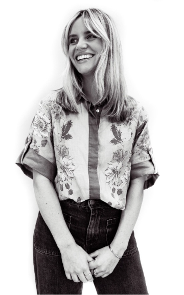 Black and white photo of a woman with shoulder-length hair smiling, wearing a short-sleeved shirt with floral and animal print details, and dark jeans.