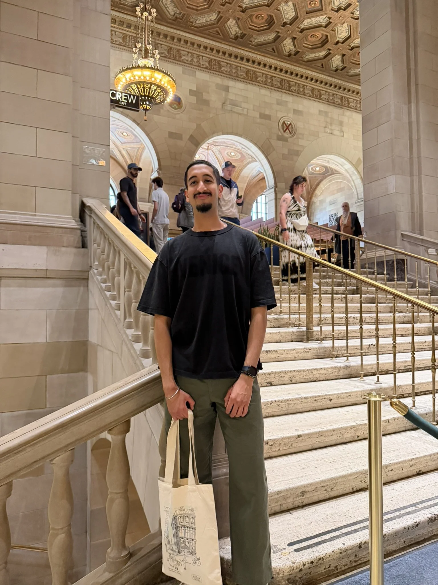 A young man with dark hair, goatee, and mustache standing on a staircase in an elegant, historic building. He is smiling, wearing a black t-shirt and olive green pants, and holding a beige tote bag with a drawing of a building. The background features ornate ceilings, arched windows, groups of people walking, and a chandelier hanging above.