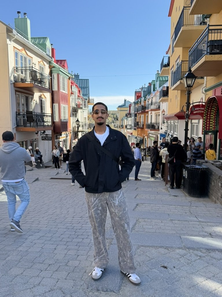 A man standing in the middle of a vibrant city street with colorful buildings, several people walking and talking, and a clear blue sky above.