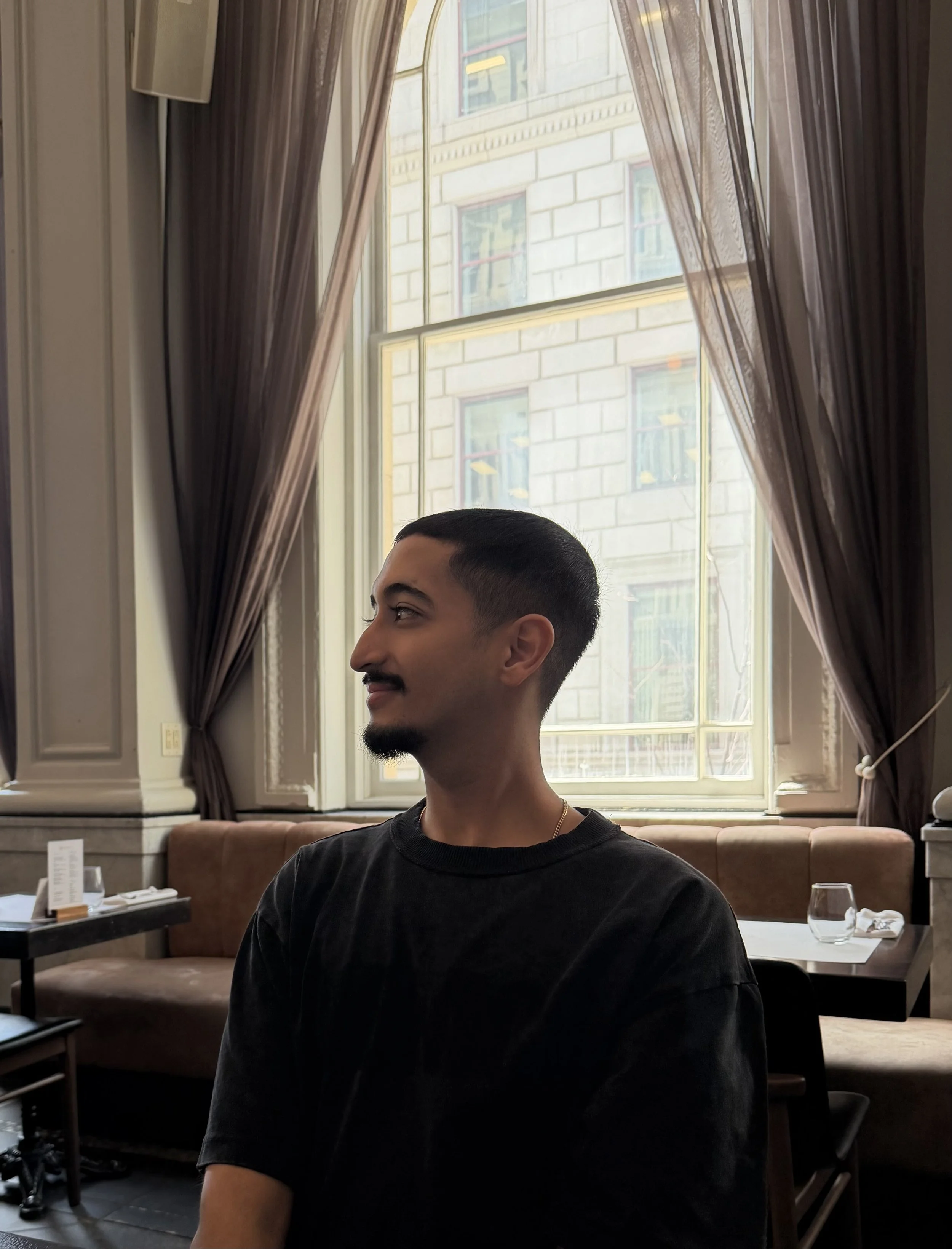 A young man with short black hair, a mustache, and a goatee, looking to the left, sitting in a restaurant with large windows, brown curtains, a beige cushioned bench, and a dark table set with glasses and utensils.