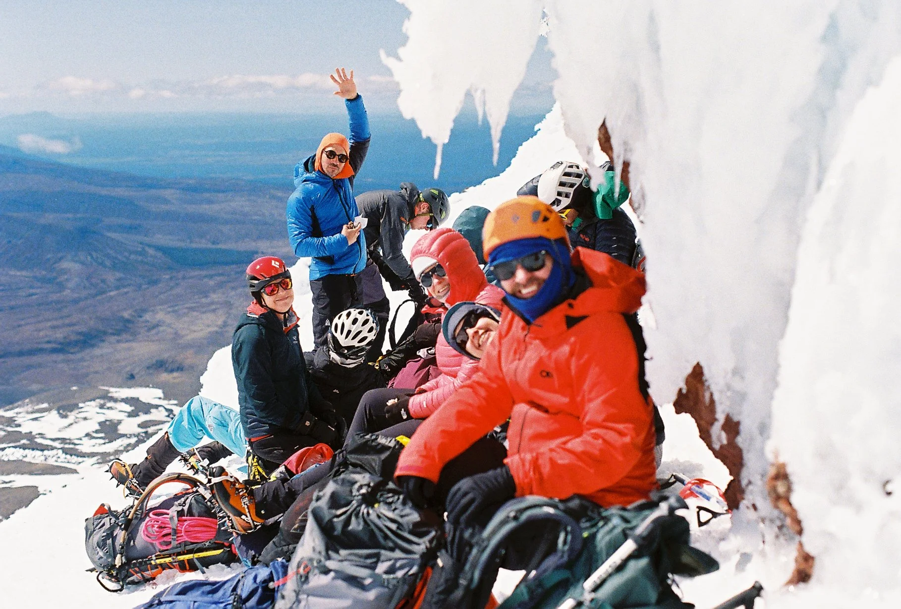 A group of climbers resting on a snowy mountain slope, wearing helmets and winter gear, with some waving and smiling at the camera.