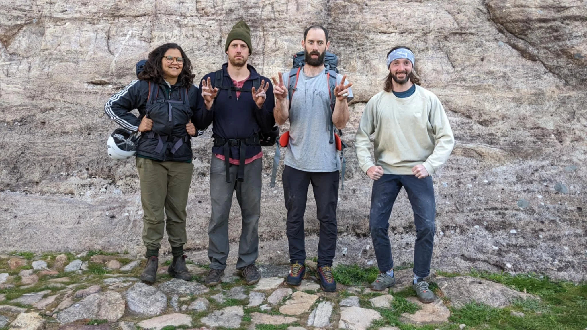 Four hikers standing in front of a rocky wall, dressed in outdoor gear, with backpacks, making peace signs or fists, smiling or looking serious.