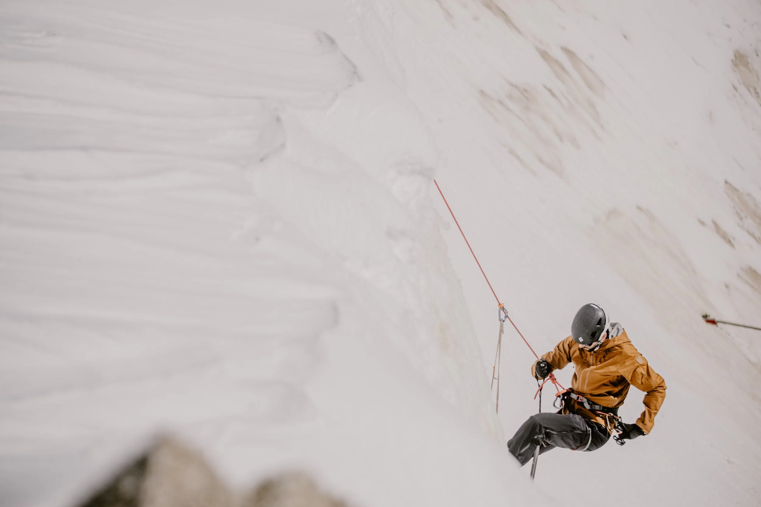 Person in an orange jacket and a helmet climbing a snow-covered mountain, using climbing gear and ropes.
