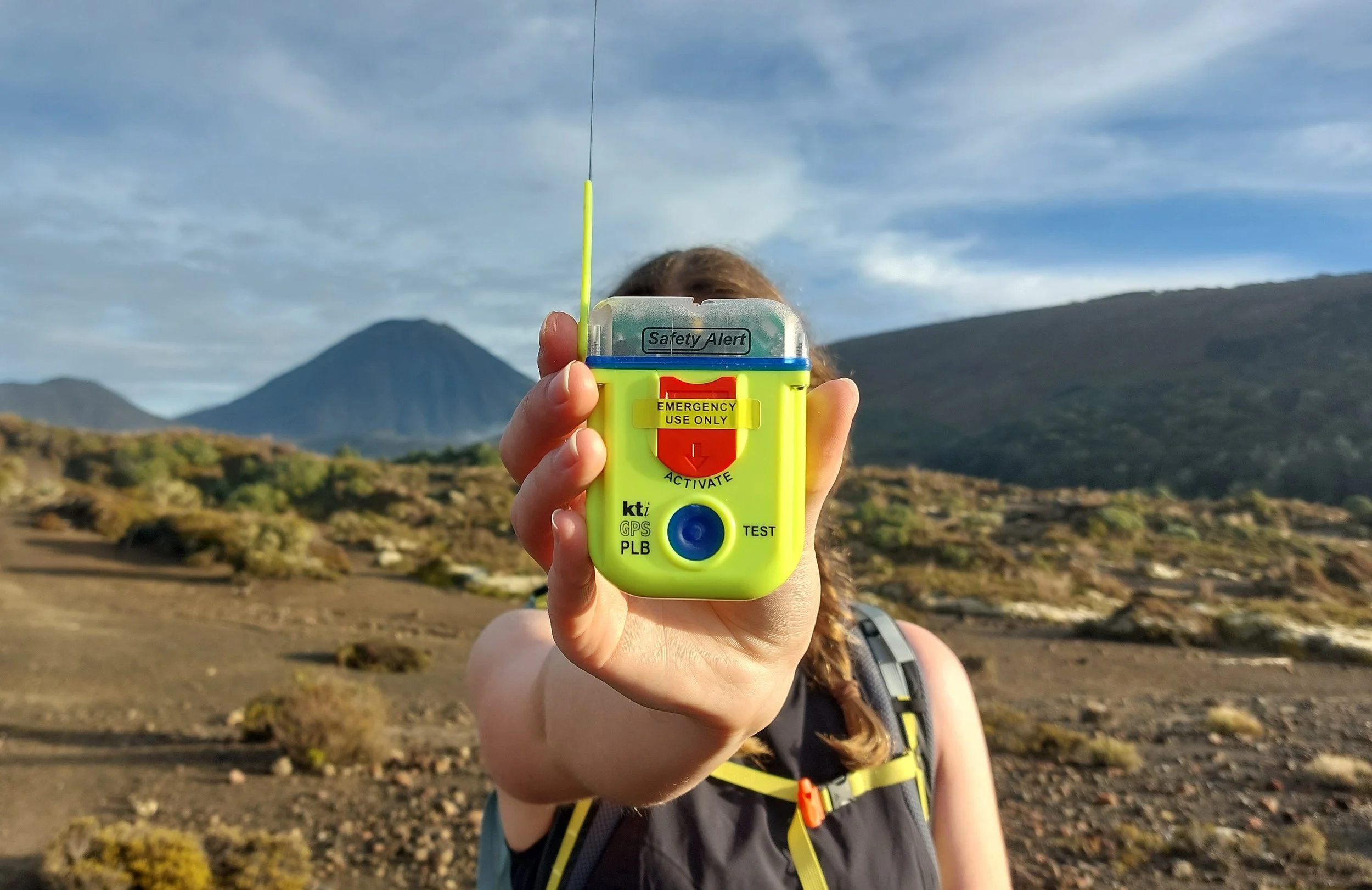 Person holding a GPS device labeled 'Safety Alert' in a mountainous outdoor area with blue sky and clouds in the background.