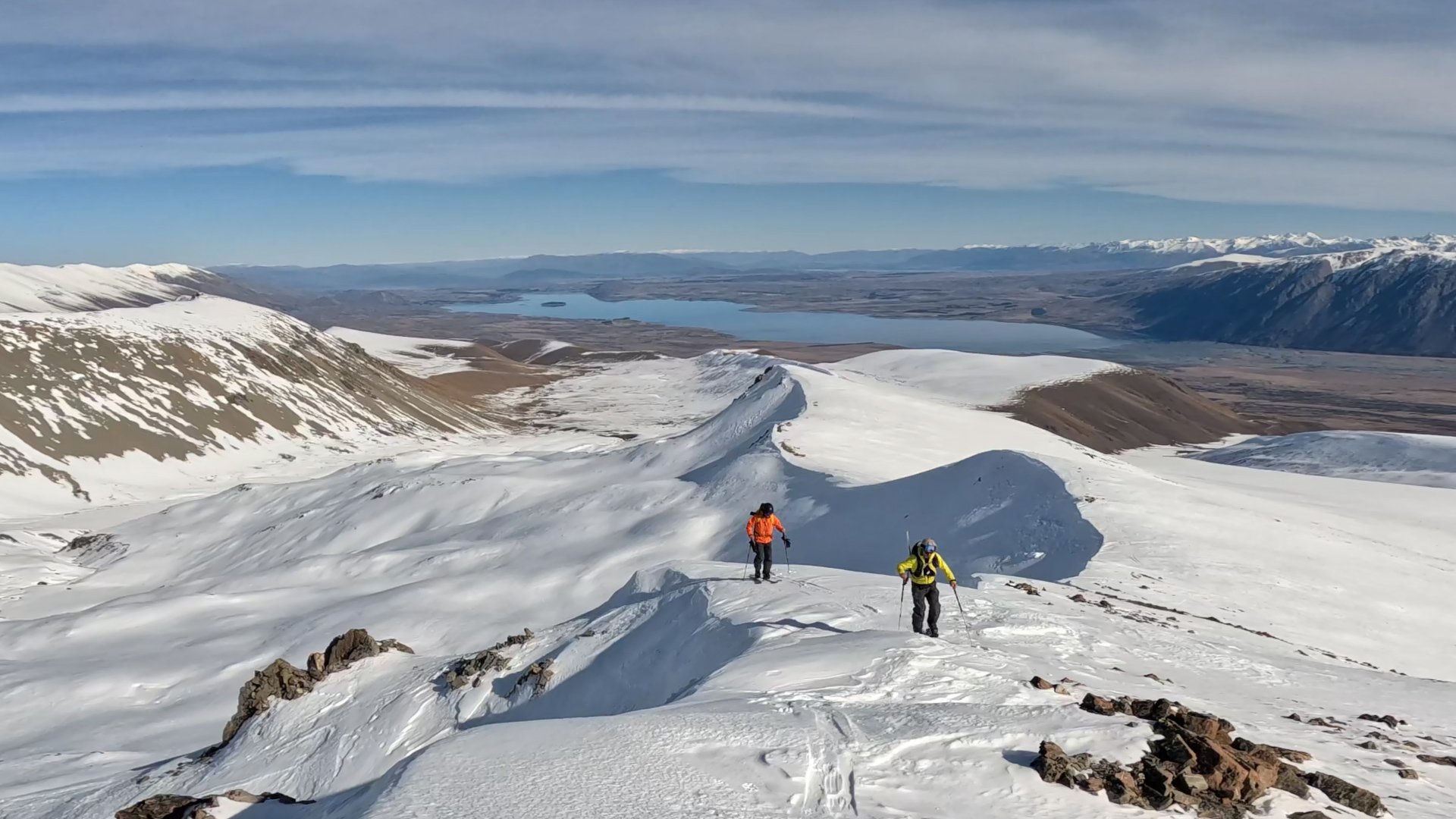 Two hikers in bright jackets trekking across a snow-covered mountain ridge with distant lakes and mountain ranges in the background.