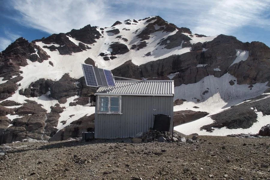 Small metal cabin with solar panels on the roof, set against a rocky, snow-covered mountain landscape.