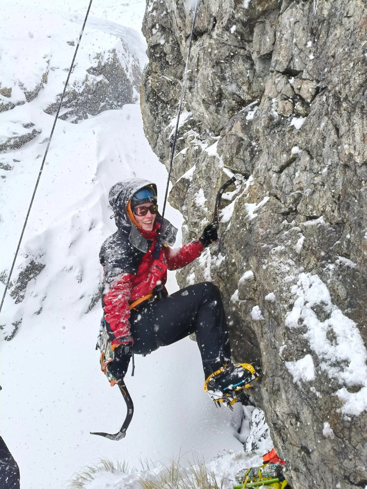 A person in winter gear, including a red and black jacket, helmet, goggles, and gloves, is climbing a snowy, rocky mountain face with climbing equipment, surrounded by snow and ice.