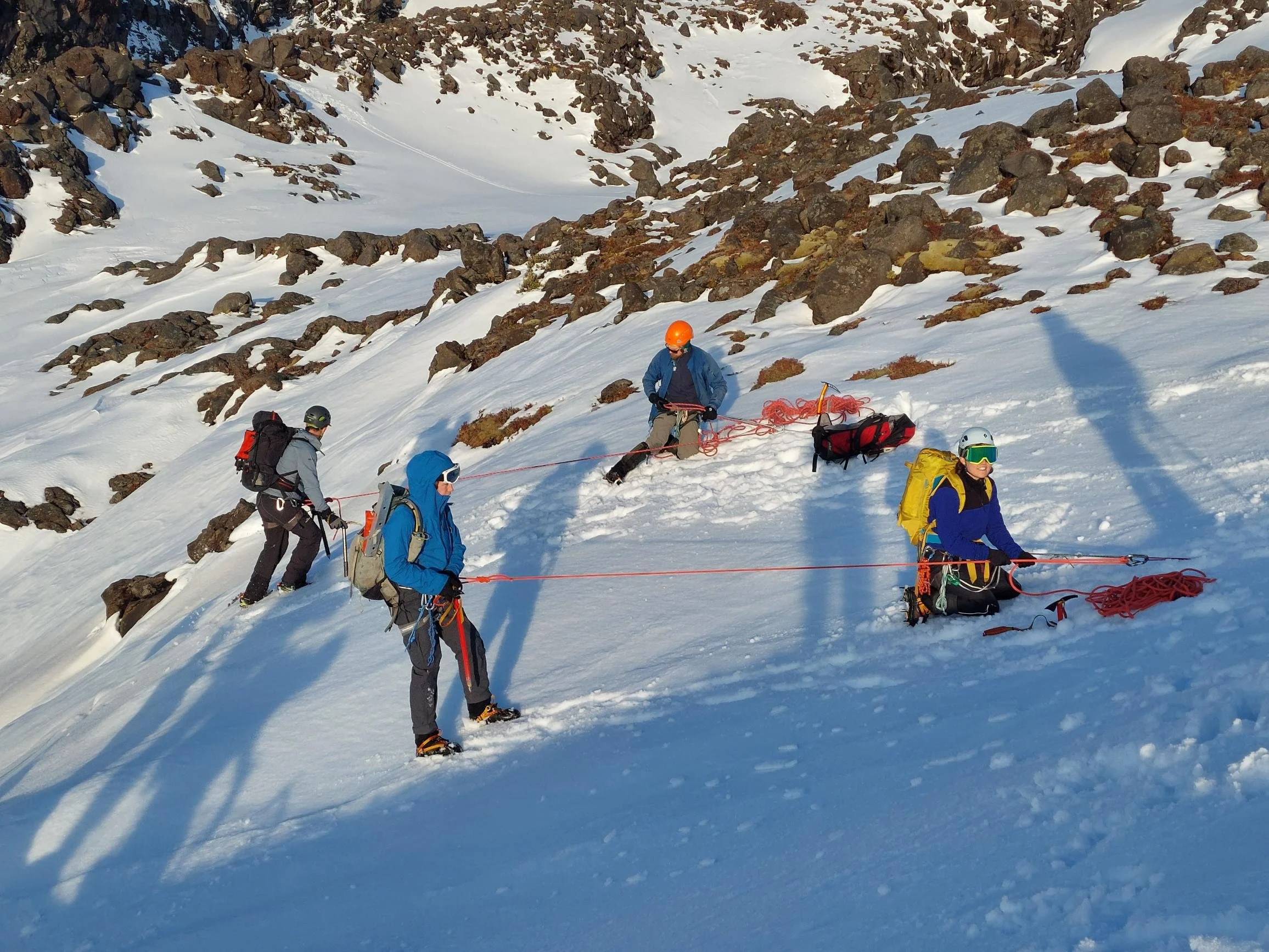 A group of people in winter gear climbing a snowy, steep mountain slope with rocks, using ropes and crampons.