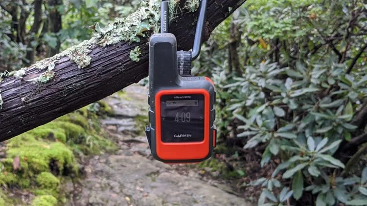 A Garmin handheld GPS device hanging from a tree branch with a carabiner, in a forest setting.