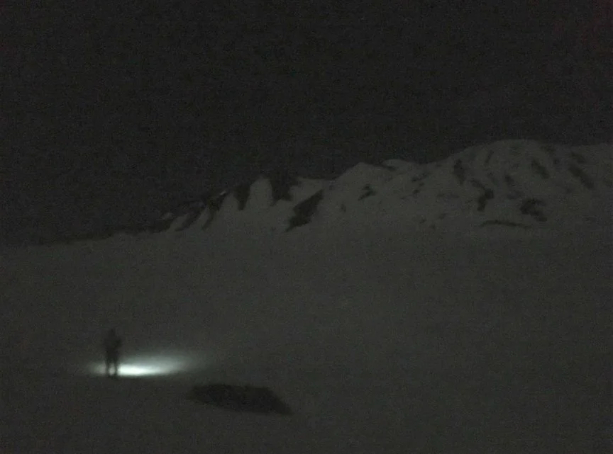 A person standing on snow-covered ground at night, holding a flashlight, with snow-capped mountains in the background.