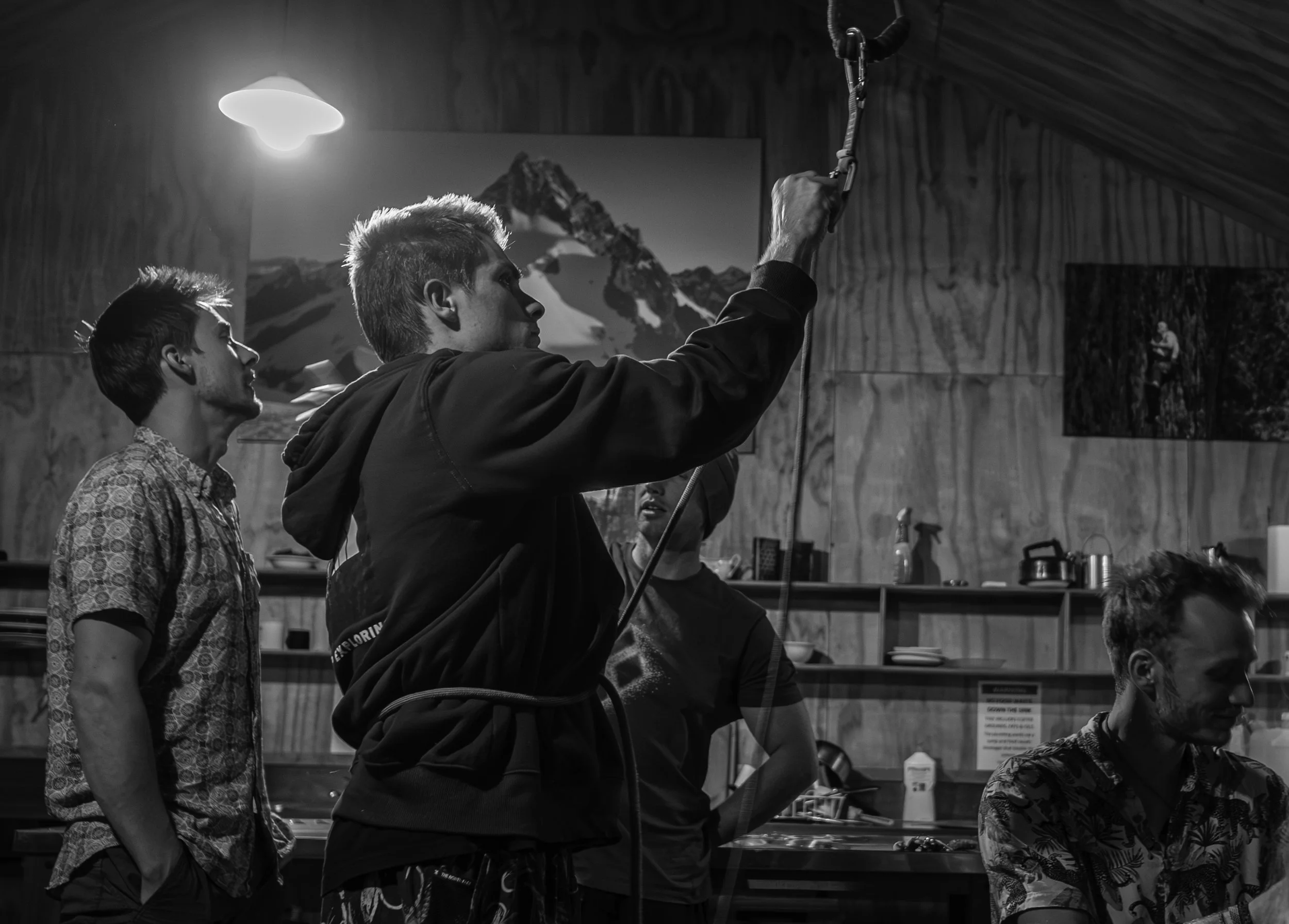 Three young men inside a climbing gym, one holding a harness and the others watching, with a mountain artwork on the wall behind them.