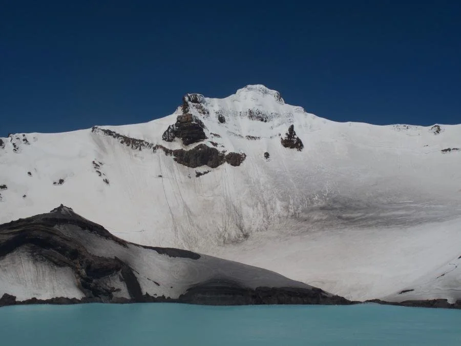 Snow-covered mountain with a glacier and a lake in the foreground under a clear blue sky.