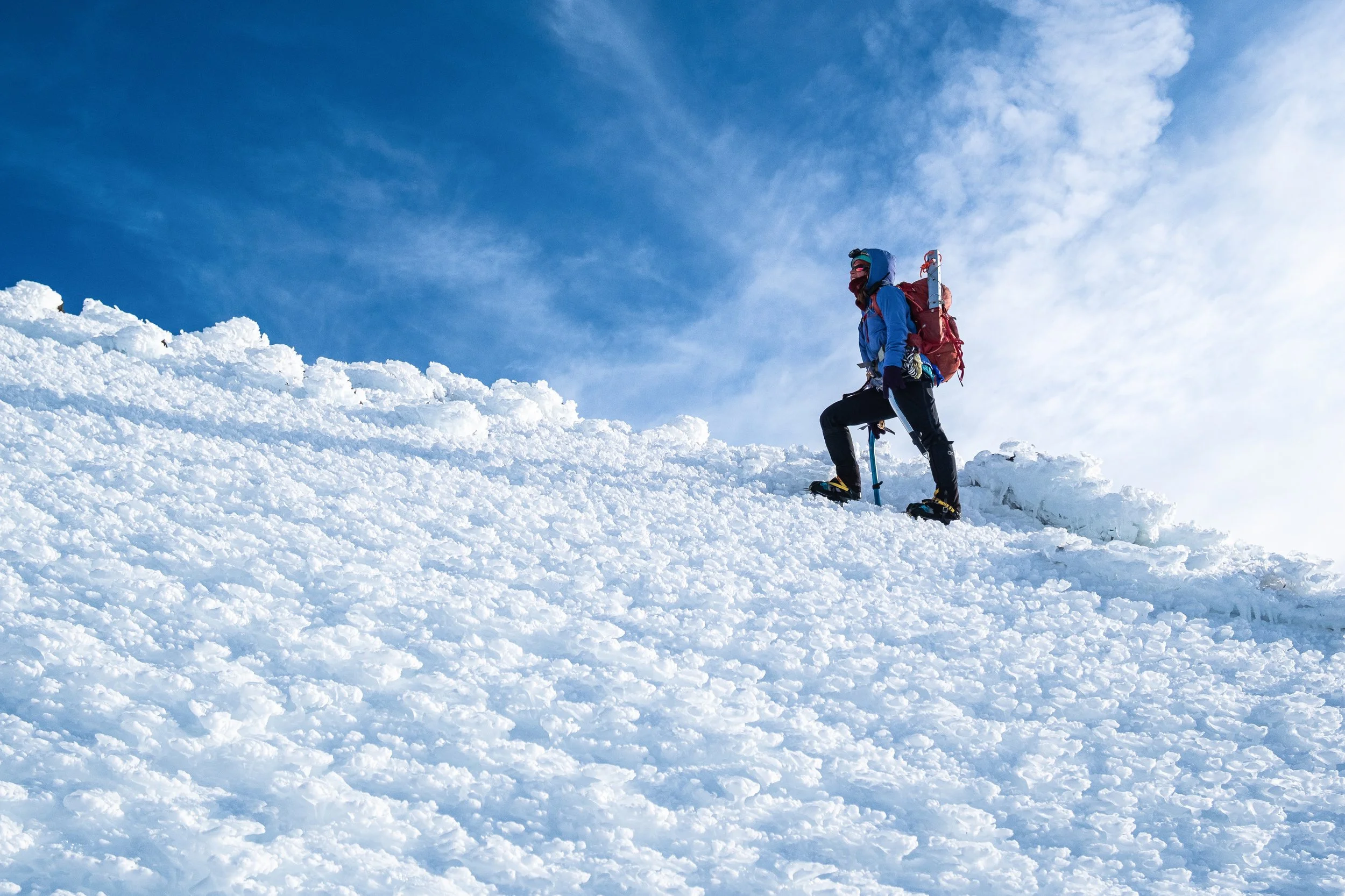 A person wearing winter gear, including a blue jacket, red backpack, and snowshoes, is standing on a snowy mountain slope under a blue sky with some clouds.