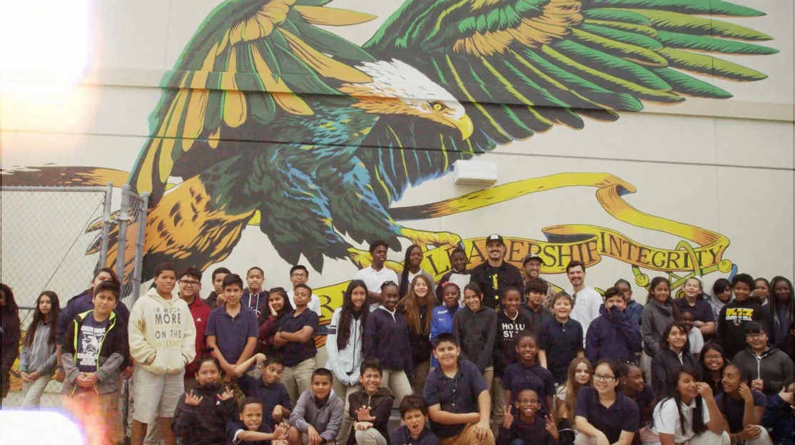 A group of students and teachers posing in front of a colorful mural of an eagle with outstretched wings. The mural includes a banner with words 'Leadership' and 'Integrity'.