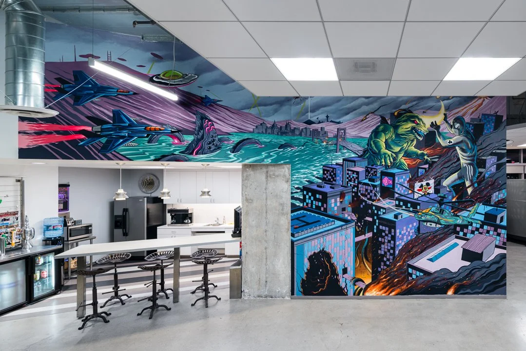 Colorful graffiti mural on a wall depicting sci-fi and monster scenes, with buildings, flying ships, a cityscape, a monster, a giant snail, a robot, and a fighter jet, in an industrial-style space with a kitchen area in the foreground.