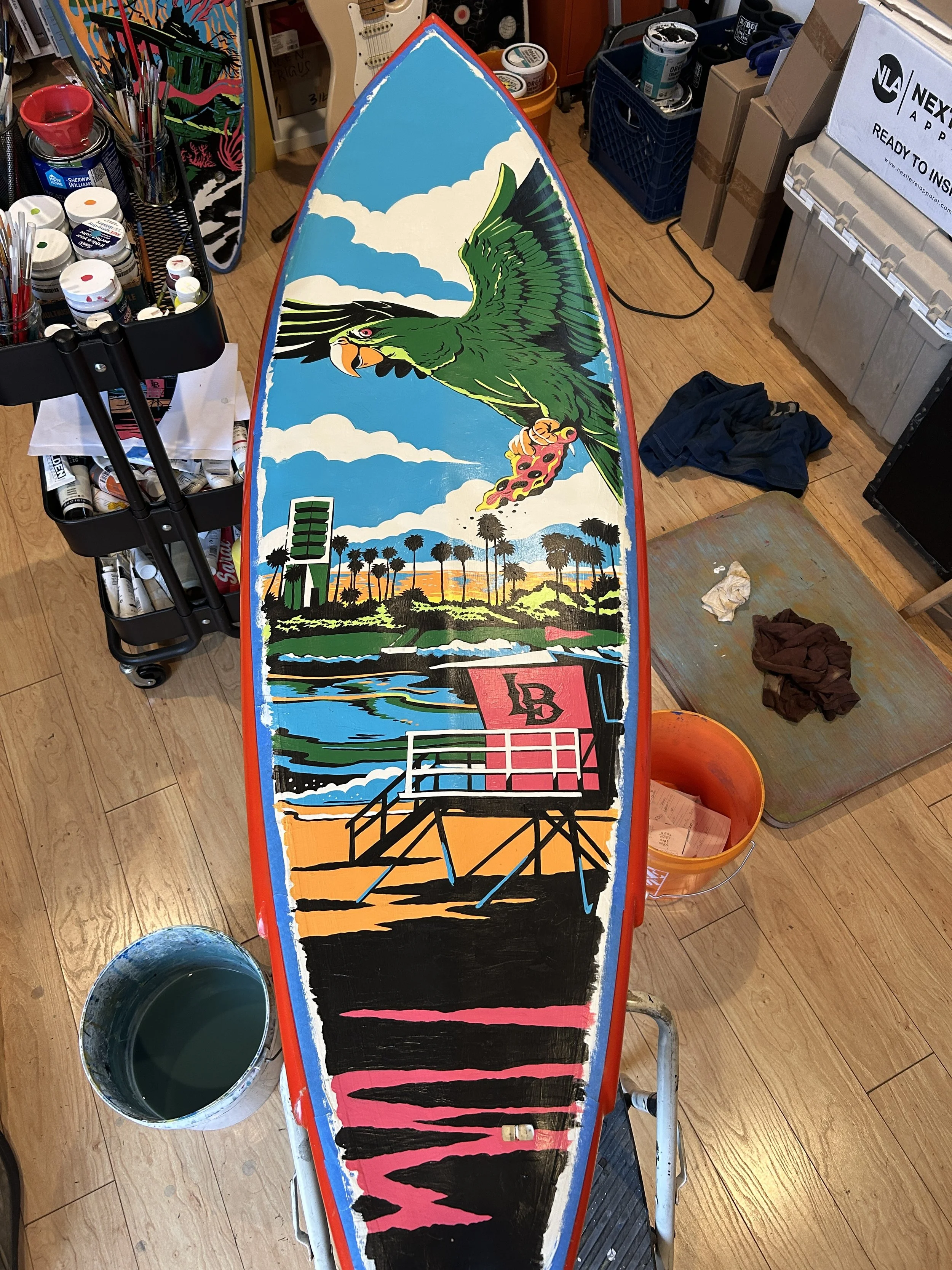 A colorful painted surfboard featuring a large green bird with yellow beak, flying over palm trees, with a bright sky background. The board is in a workshop surrounded by various paint supplies and tools.