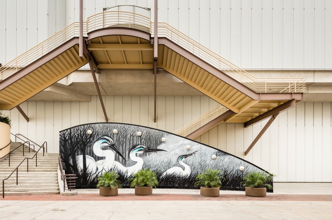 Mural of three herons among tall grass with a forest background, located beneath outdoor stairs on an urban building exterior.