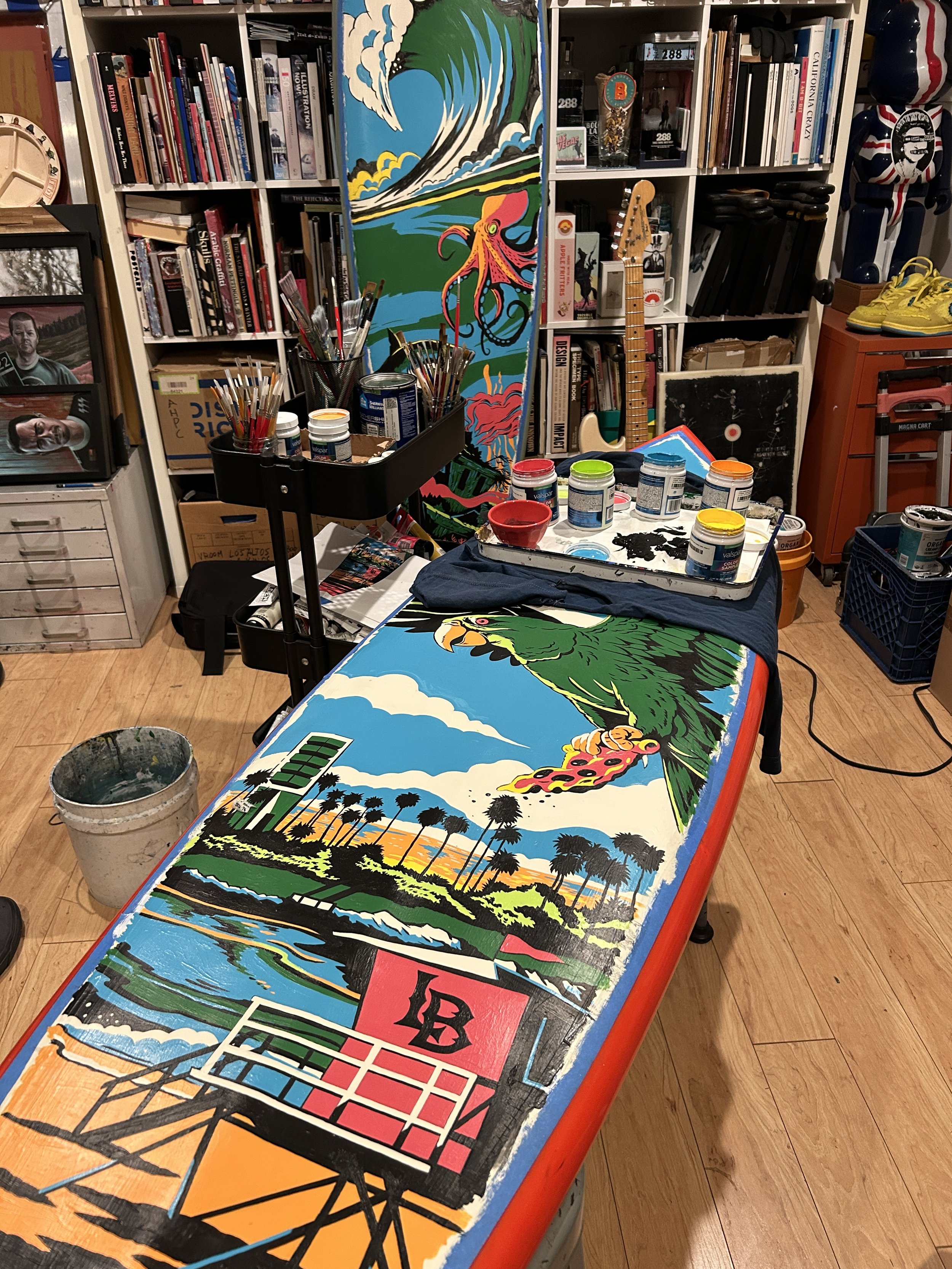 A workspace with art supplies and three uncompleted colorful paintings on a table. The paintings depict a landscape with sunset, palm trees, a billboard, and a parrot with a cityscape background.
