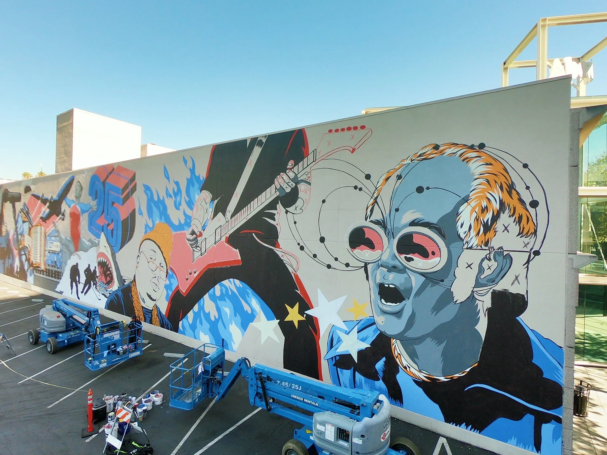 A large colorful mural on an exterior wall features a stylized illustration of a person with sunglasses and a bear, along with abstract shapes and designs. There are construction lift platforms and painting supplies in the parking lot below the mural.