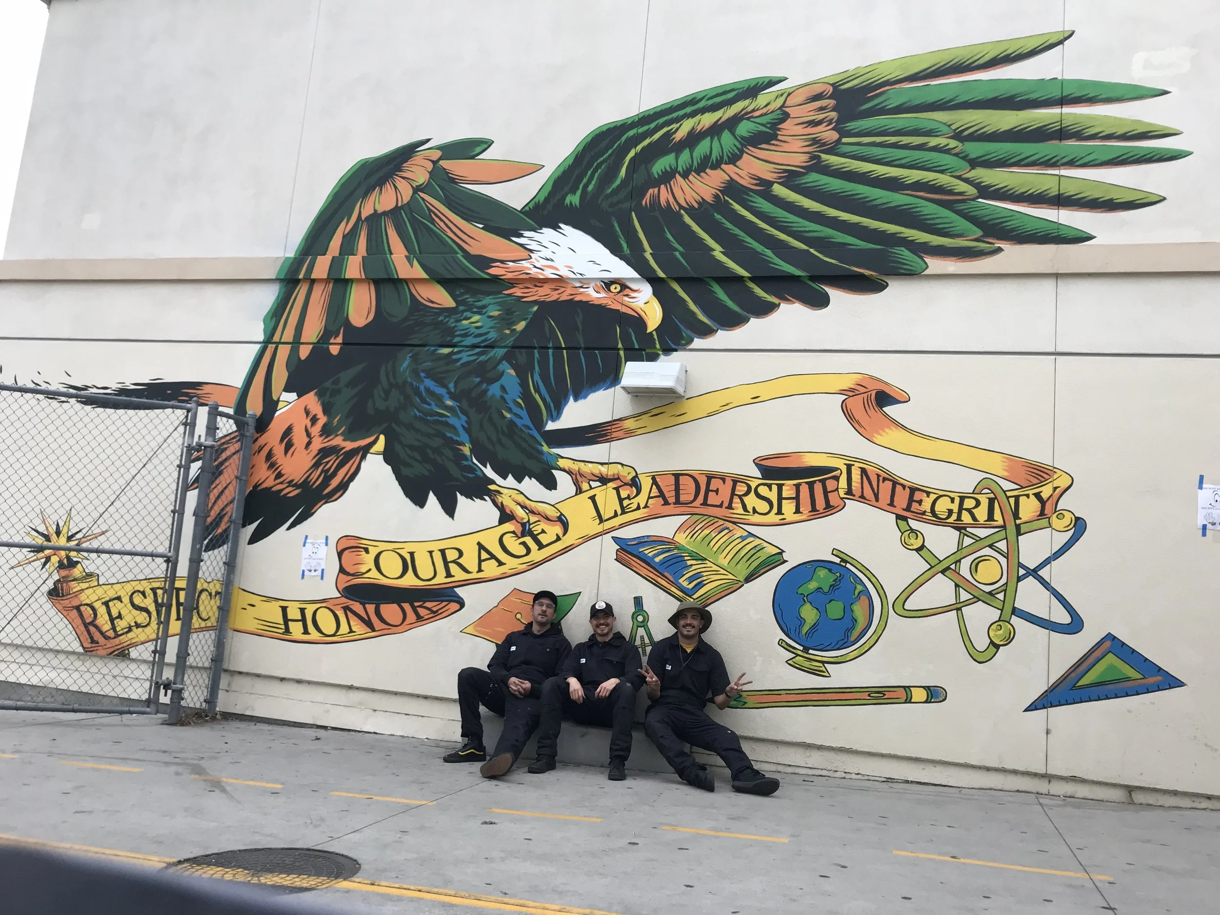 A colorful mural of a soaring eagle on a wall, with three people sitting in front of it, making peace signs with their hands.