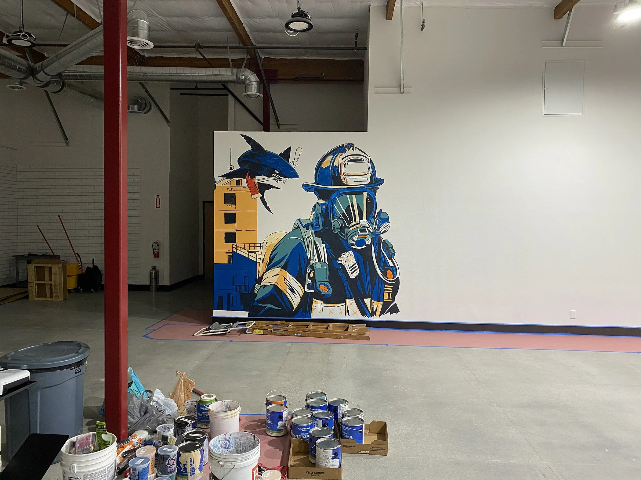 Art supplies and painting equipment in a large indoor space with a painted mural of a scuba diver and a shark on the wall.