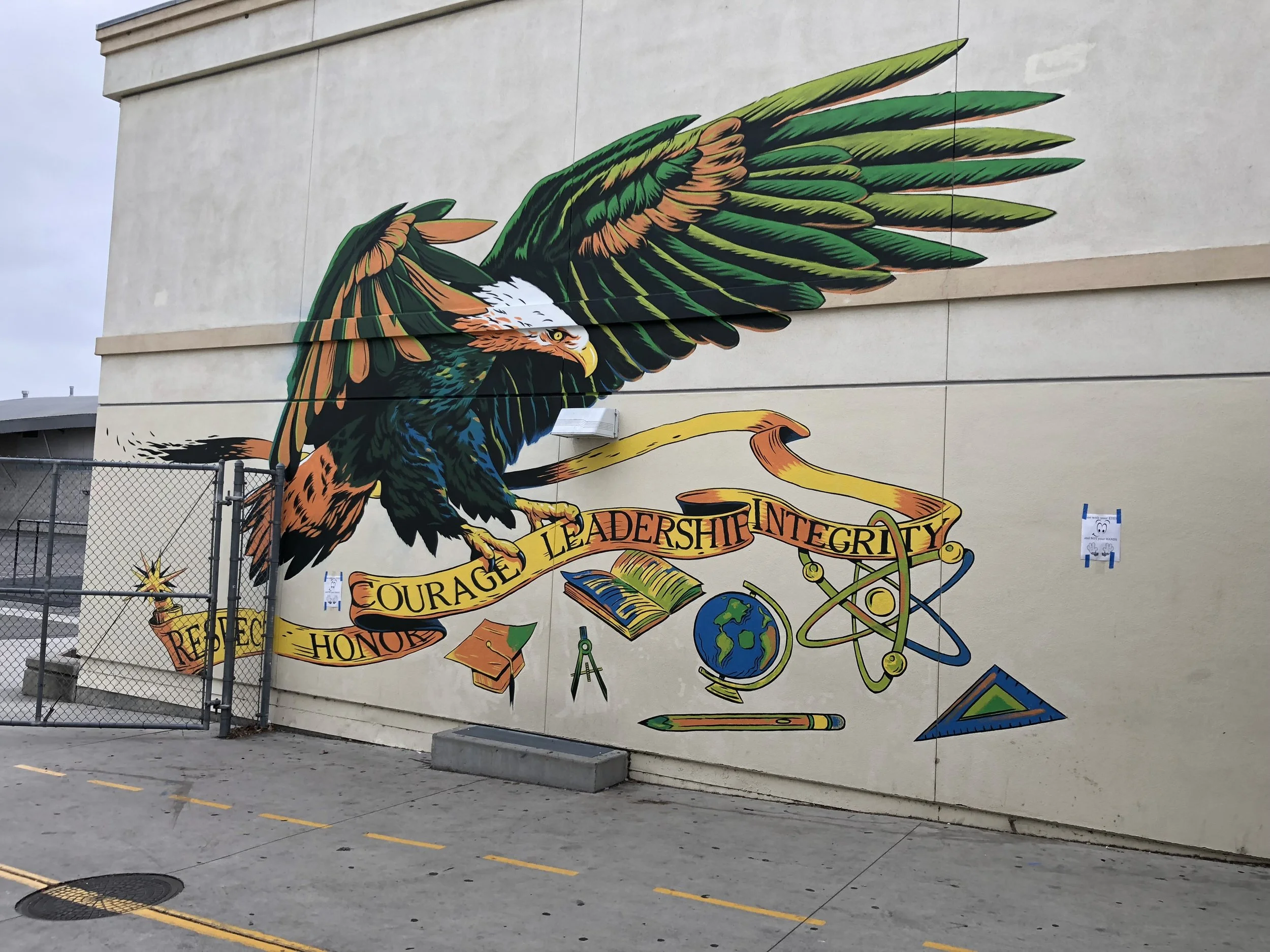 A colorful mural depicts a fierce eagle with outstretched wings on a building wall, surrounded by images of books, a globe, and science symbols, with words like 'Courage,' 'Integrity,' and 'Respect' written on a ribbon.