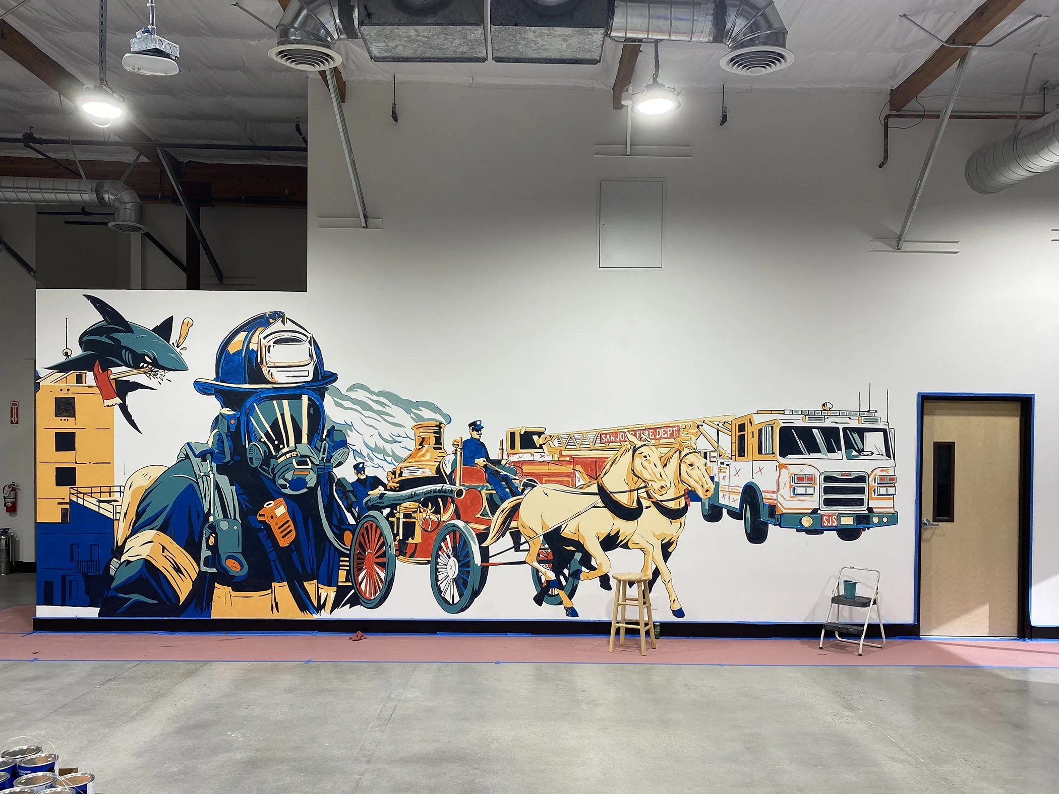 A large colorful mural in progress depicting emergency vehicles, firefighters, and equipment with a white background, located in an indoor space with visible ceiling ductwork and lighting.