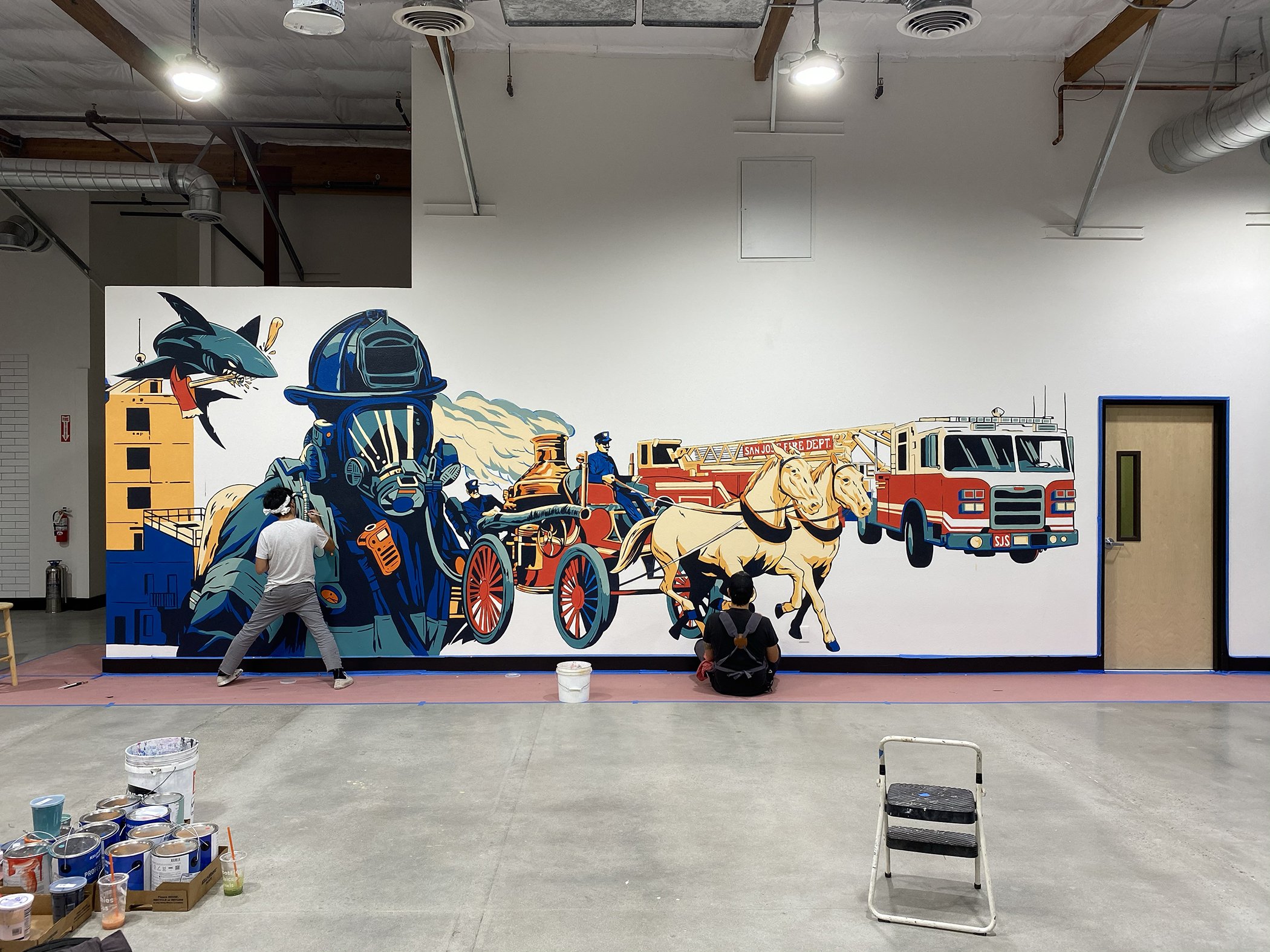 A large colorful mural depicting fire fighters, fire trucks, horses, and a sea creature with a fire hose on a white wall in an industrial-style room. Two people are working on the mural, with paint cans and supplies placed on the floor in front.