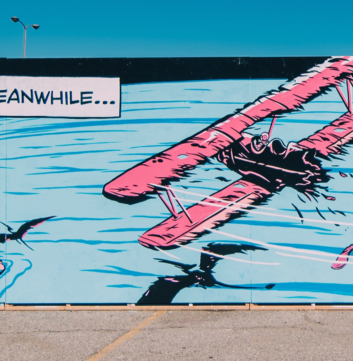 A mural painting of a pink biplane flying over water with the word 'Meanwhile...' in a white box in the top left corner.