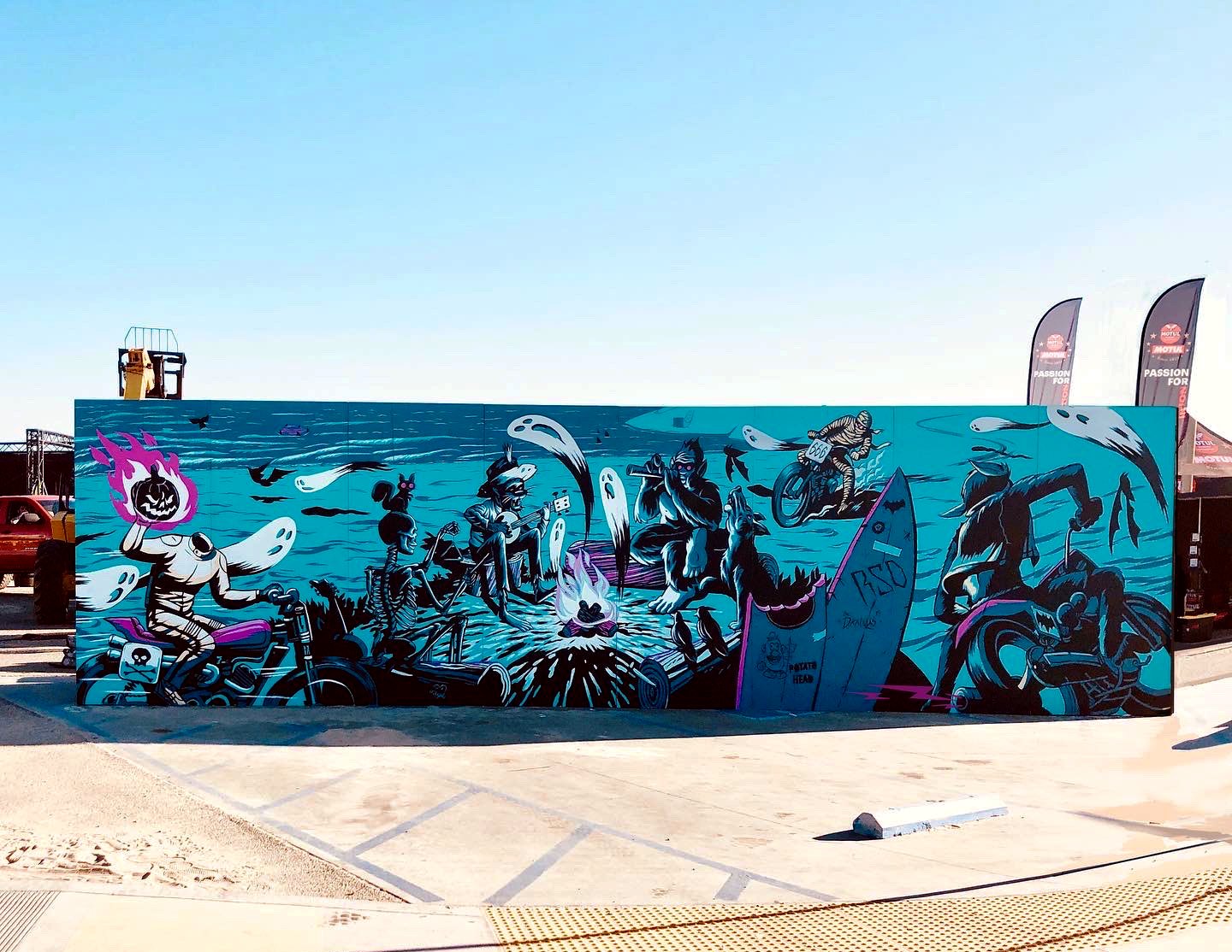 Colorful mural graffiti art with skeletons riding motorcycles and surfing, including a figure with a flaming pumpkin head, set against a bright blue background with birds and ocean waves.
