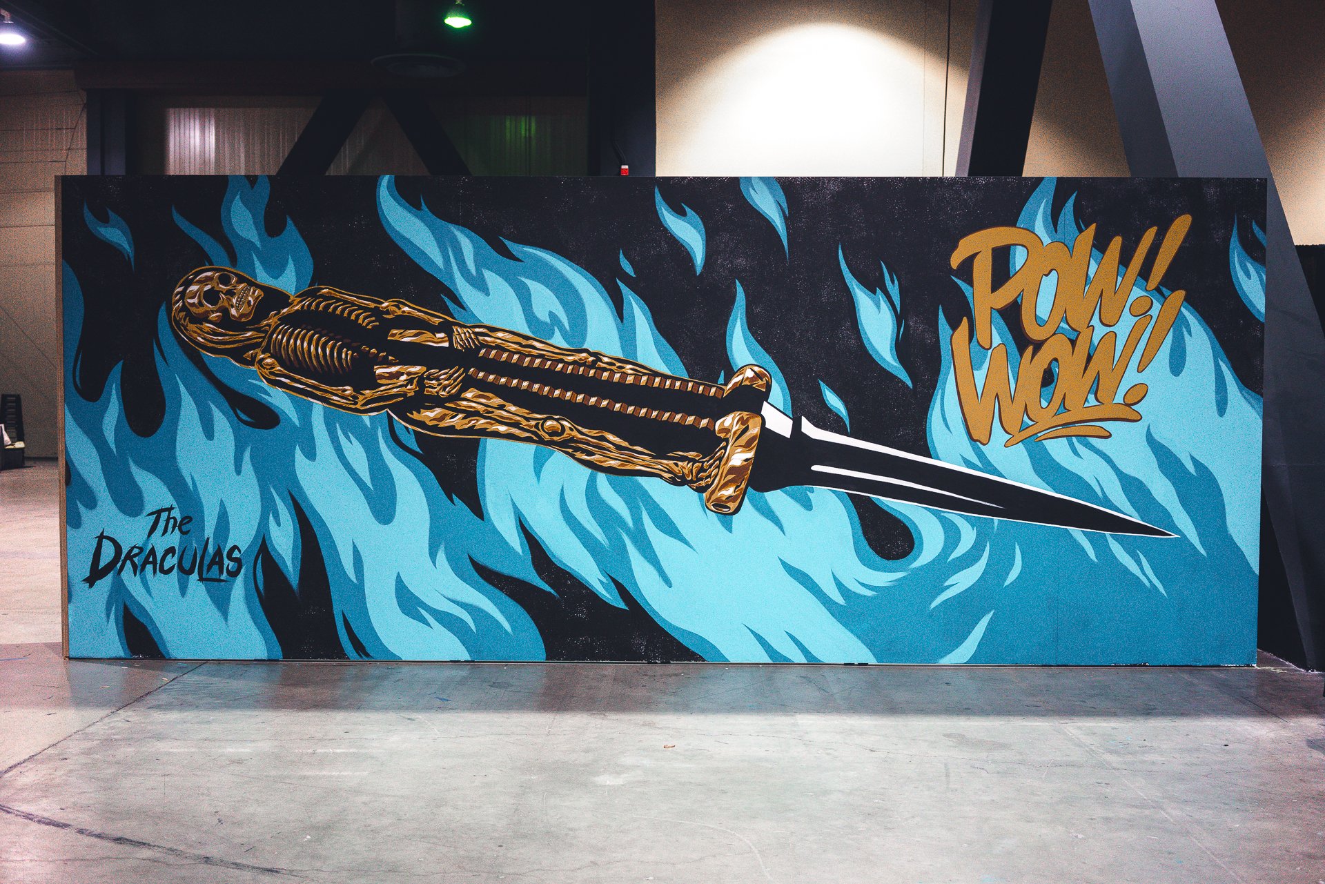 A colorful poster depicting a skeleton holding a dagger, with blue flames in the background and the words 'POW! WOW!' in bold letters. The poster is titled 'The Draculas'.