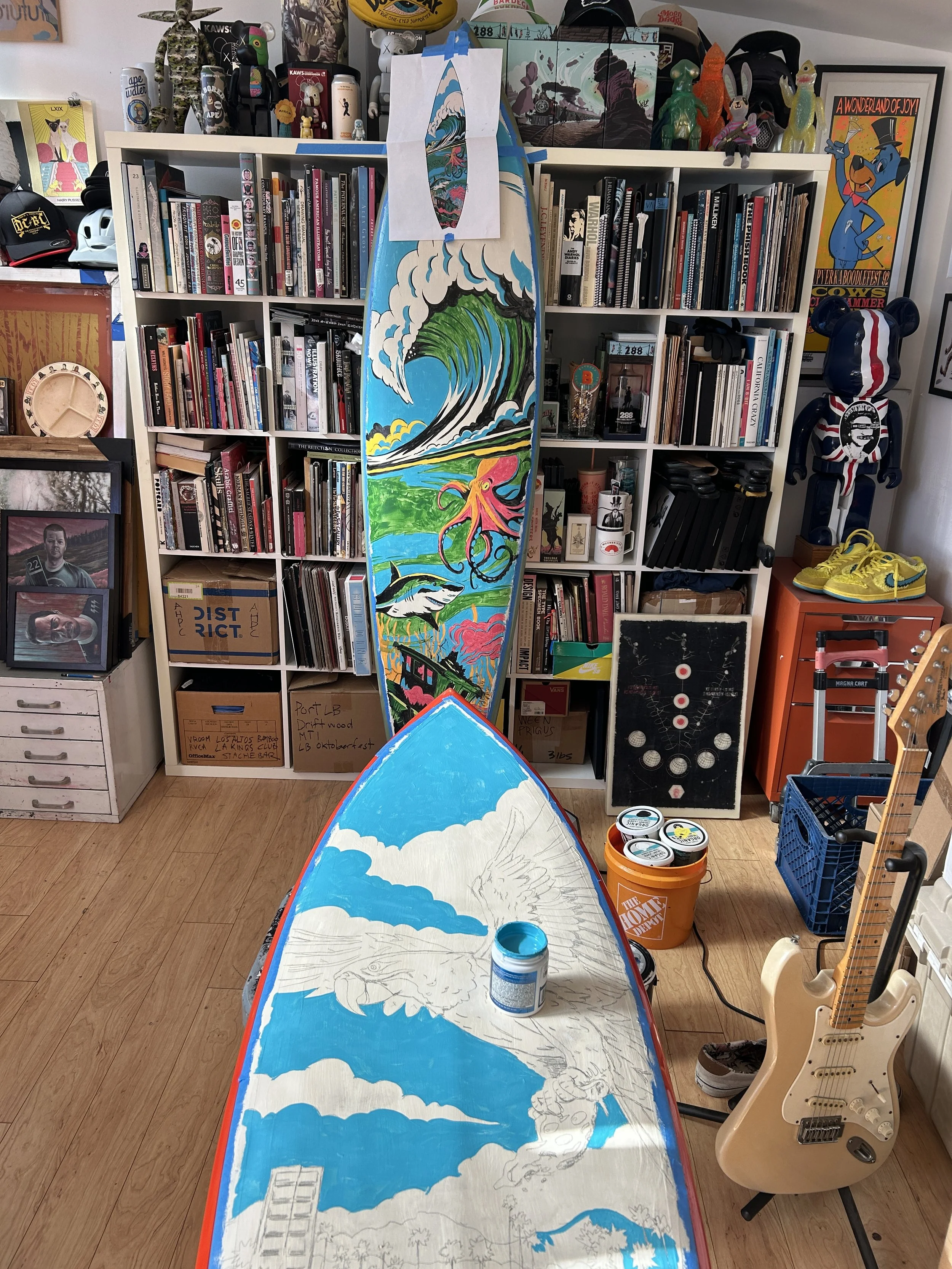 A cluttered room with bookshelves filled with books, artwork, and collectibles. In the foreground, a boat-shaped surface is being painted with a colorful landscape, including a large wave, an octopus, and other scenery. There is a Fender electric guitar on a stand, a small orange crate, and various art supplies and boxes scattered around.