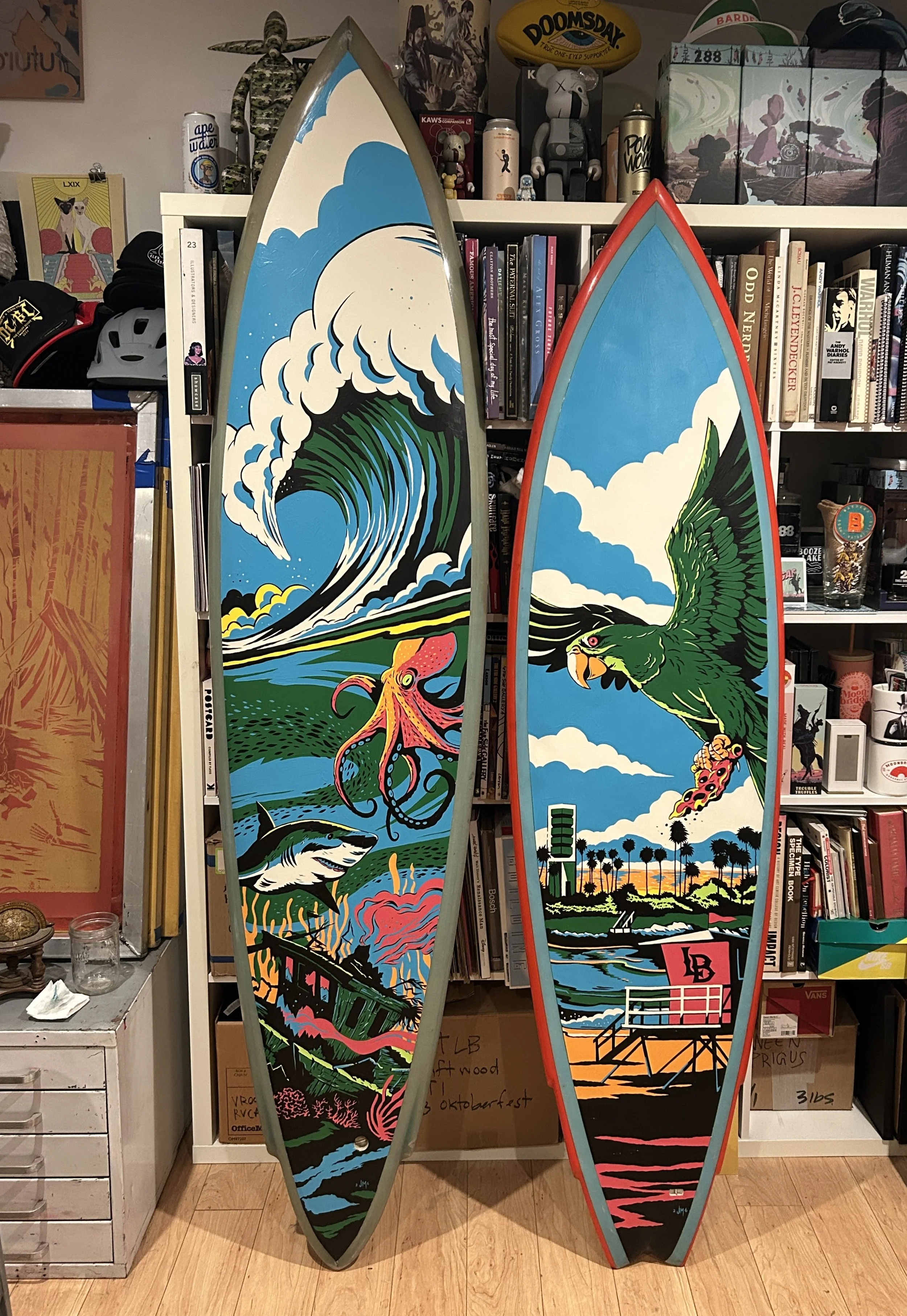 Two colorful surfboards with artistic designs leaning against a bookshelf filled with books and collectibles. The surfboard on the left features a large wave crashing with a pink octopus and a shark, while the one on the right displays a green parrot flying over a sunset landscape with palm trees.