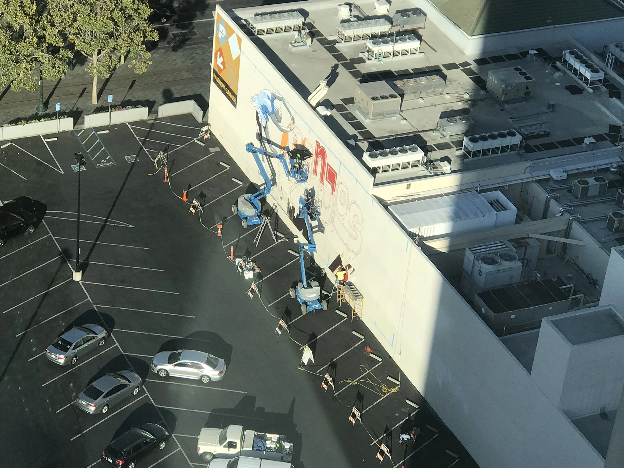 An aerial view of a parking lot with multiple parked cars and a building. A worker on a lift is painting or fixing a wall on the side of the building, with safety cones and equipment around. The building's roof has HVAC units and other mechanical equipment.