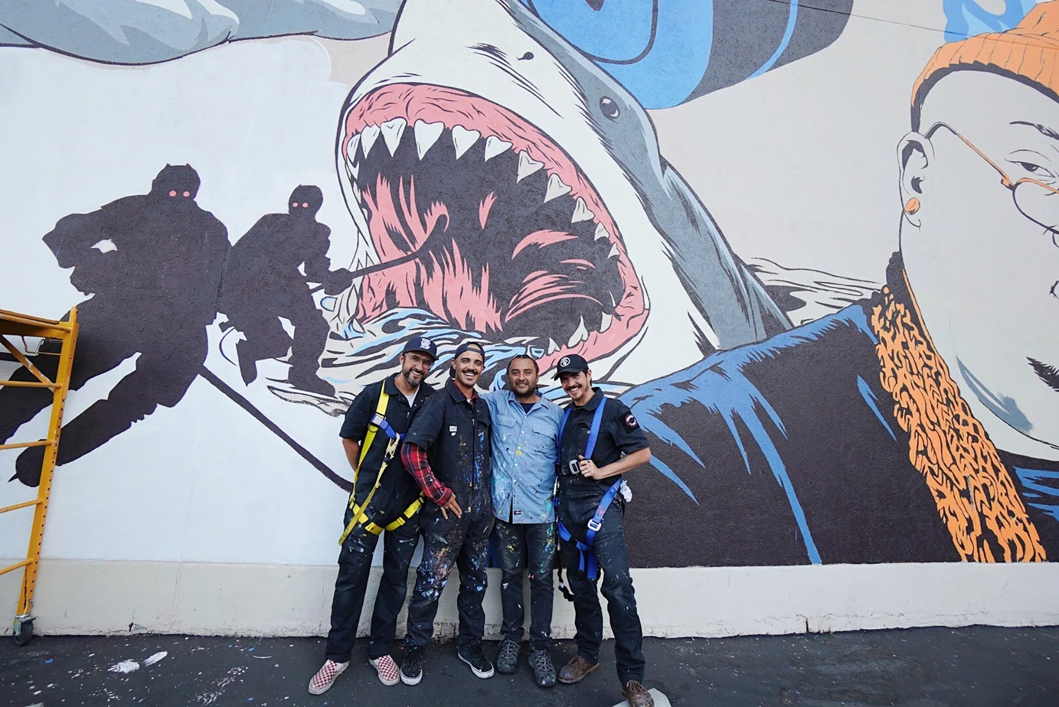 Four men standing in front of a colorful mural of a shark with its mouth open, on a wall. The men are smiling and wearing casual clothing and painting gear.