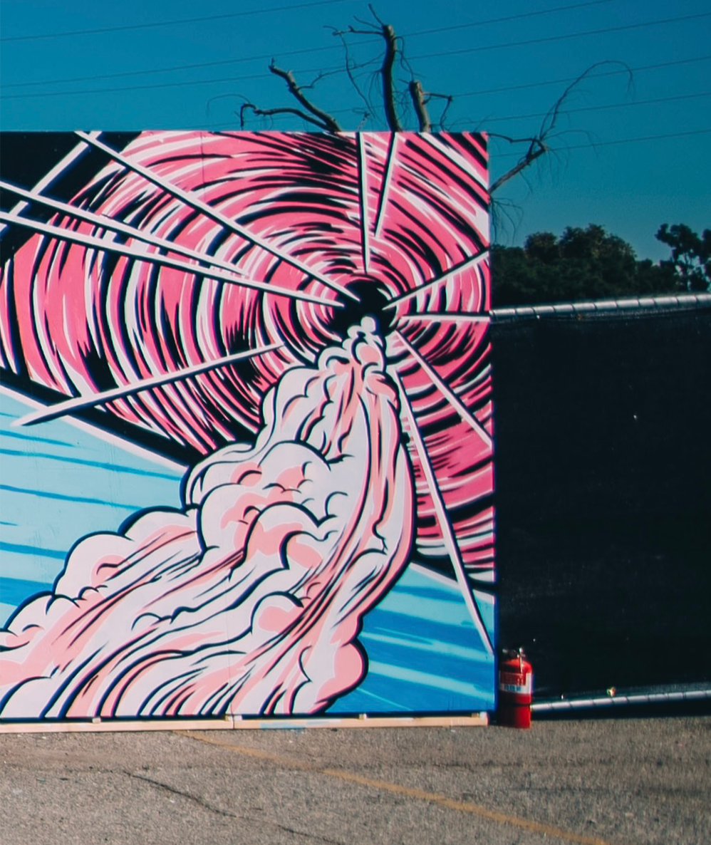 Street art mural of a stylized pink and black tornado with a hand reaching into the vortex, set against blue sky with power lines and a leafless tree in the background.