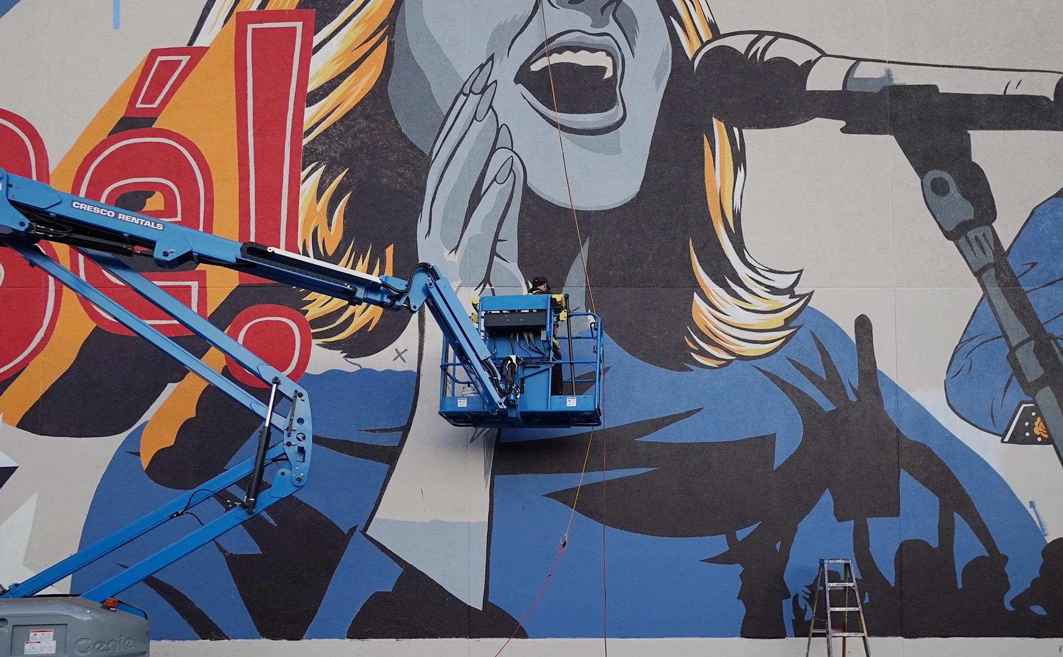 A large mural of a woman with long blonde hair, her mouth open as if shouting or singing, with her hand holding a microphone and her other hand near her face. There are two blue cherry pickers in front of the mural, one with a worker on it near the bottom left and another with a worker on it at the center, painting parts of the mural.