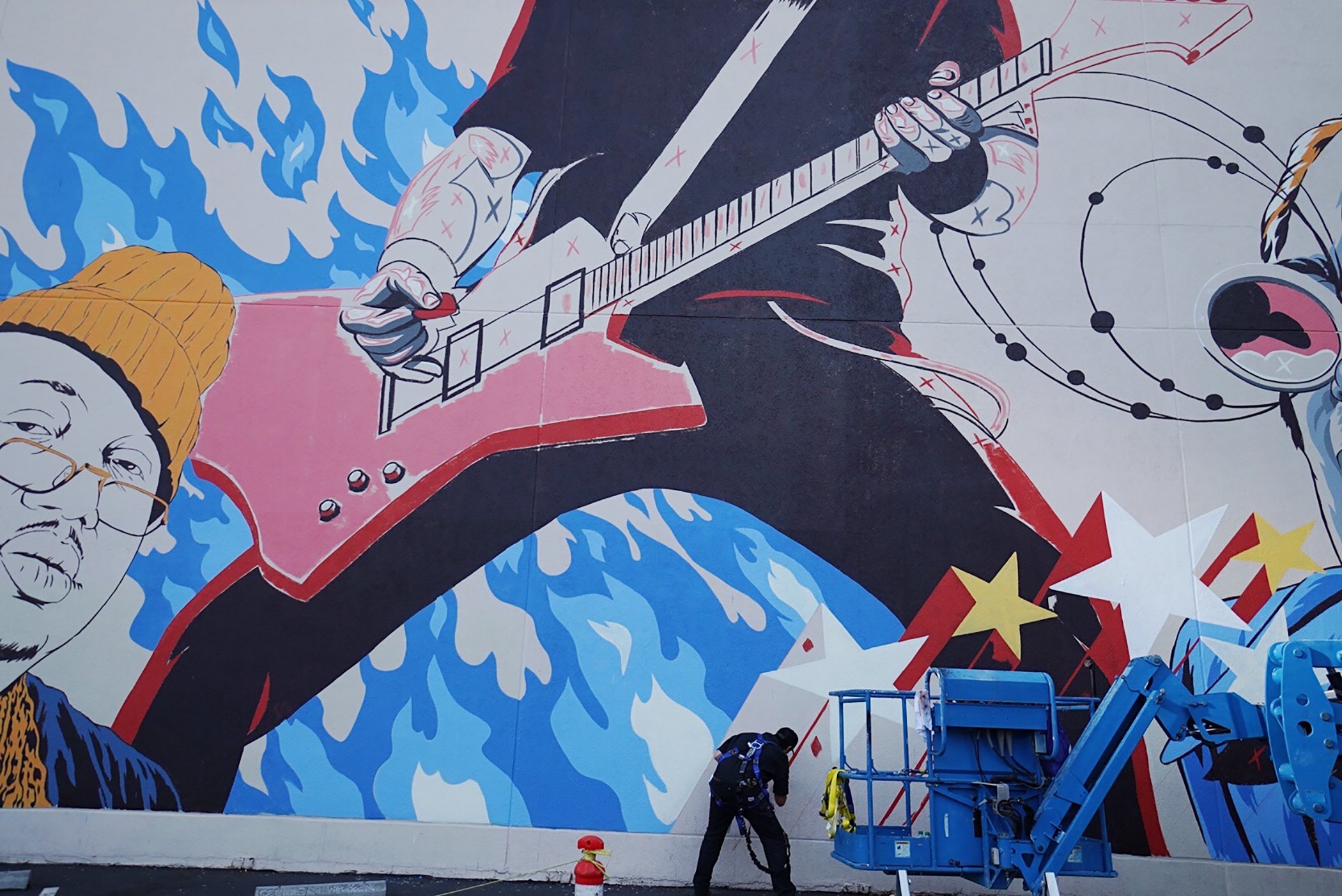 A mural of a person playing an electric guitar, wearing glasses and a yellow beanie, with a background of blue flames and abstract designs. Two workers are installing or maintaining the mural using a cherry picker.