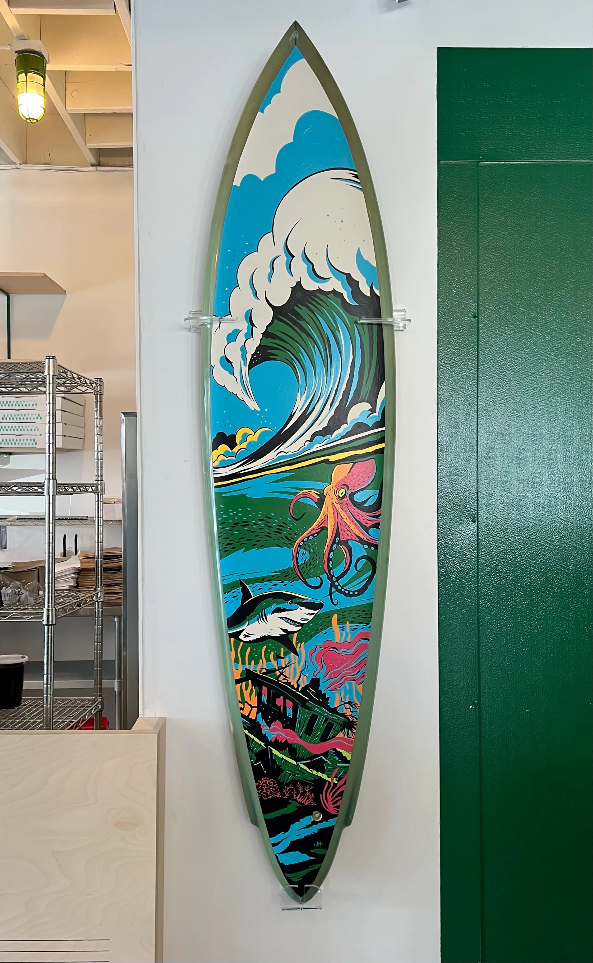 Colorful surfboard with a vibrant ocean scene painting, including a large breaking wave, a pink octopus, a shark, a lighthouse, and a pink coral reef, mounted on a white wall.