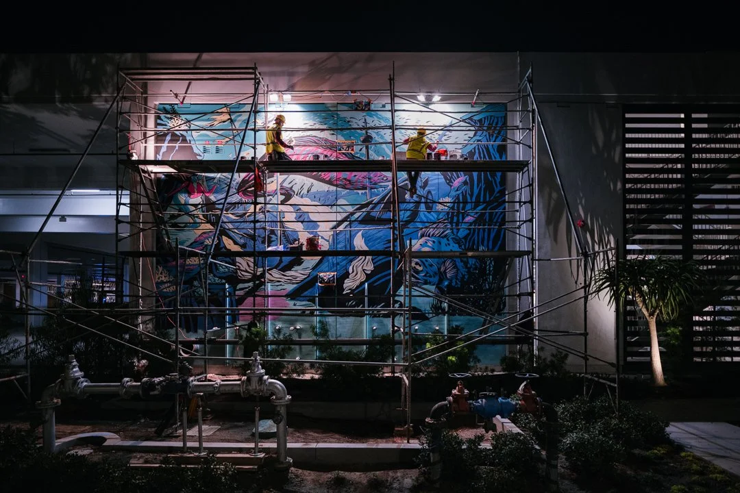 Workers on scaffolding painting a large, colorful mural on a building wall at night. The mural depicts abstract and stylized images of characters and nature, with vivid blue, pink, and purple tones.