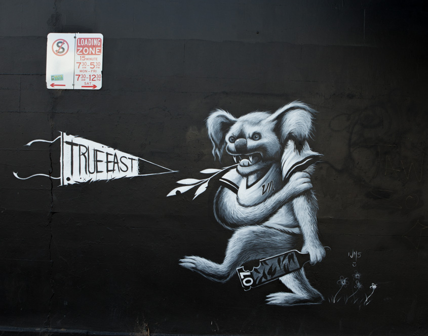 Street graffiti of a koala with a friendly expression, wearing a shirt, holding a flag that reads 'TRUE EAST', and carrying a book labeled 'LOM'. The background is black with a parking sign above.